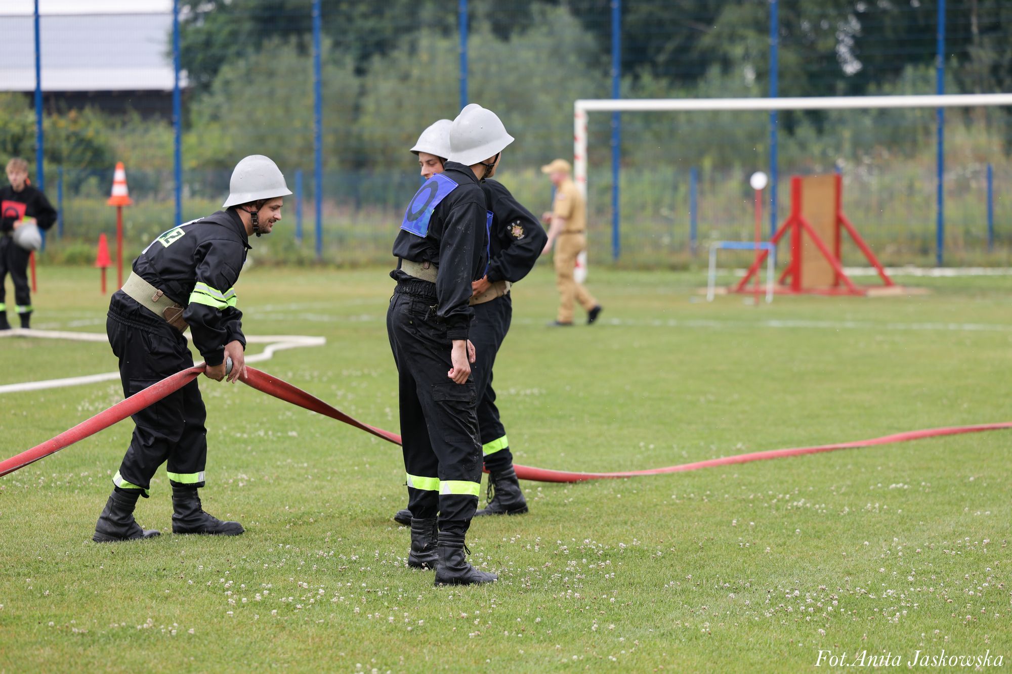 Czterech mężczyzn w czarnych mundurach i białych hełmach trzyma czerwony wąż strażacki na zielonym boisku sportowym. W tle piłkarska bramka i sprzęt treningowy.