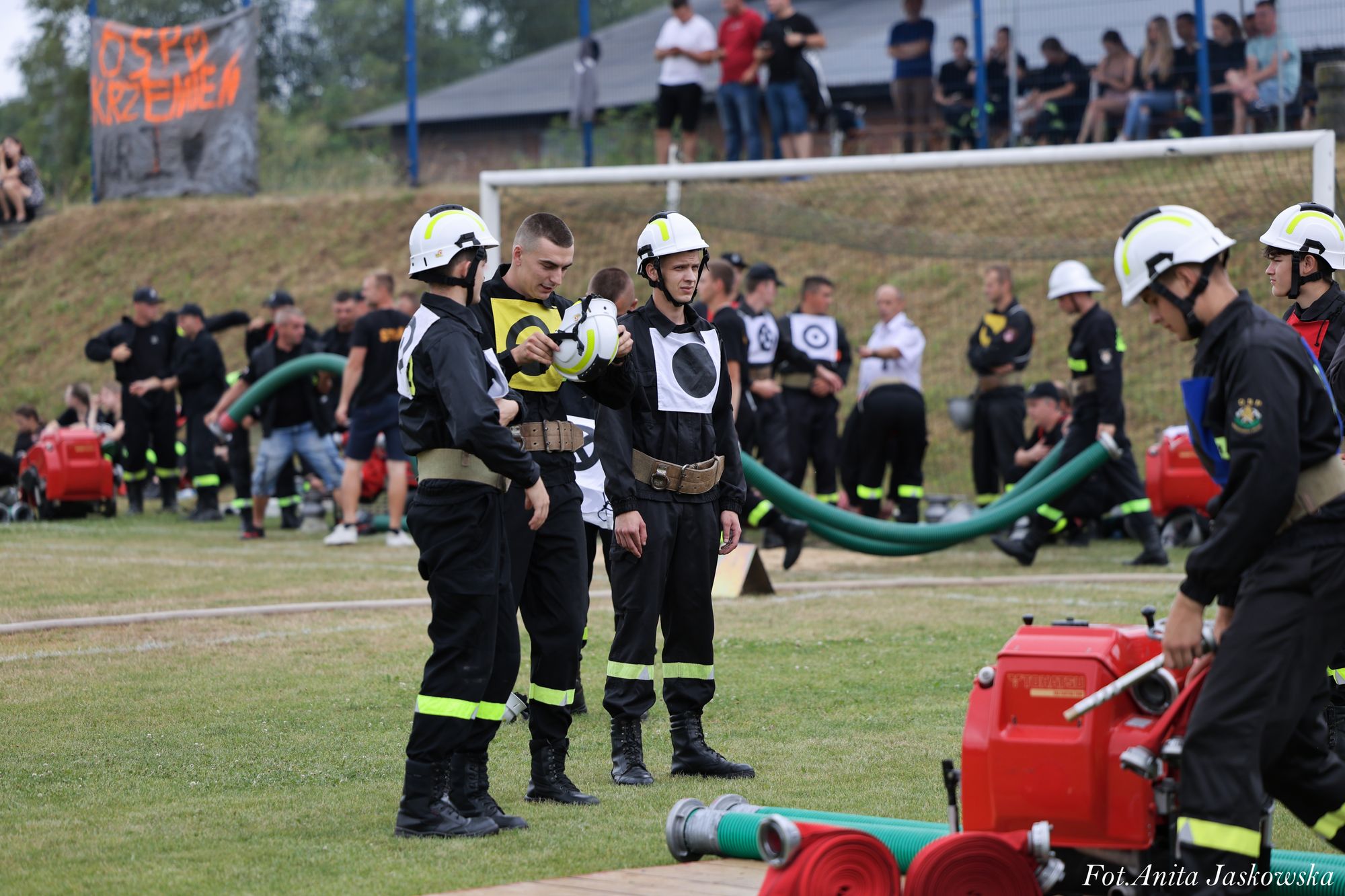 Grupa młodych strażaków w czarnych mundurach z odblaskowymi paskami i białymi hełmami stoi na trawie, w tle sprzęt strażacki, zielone i czerwone węże, oraz widownia na pagórku.