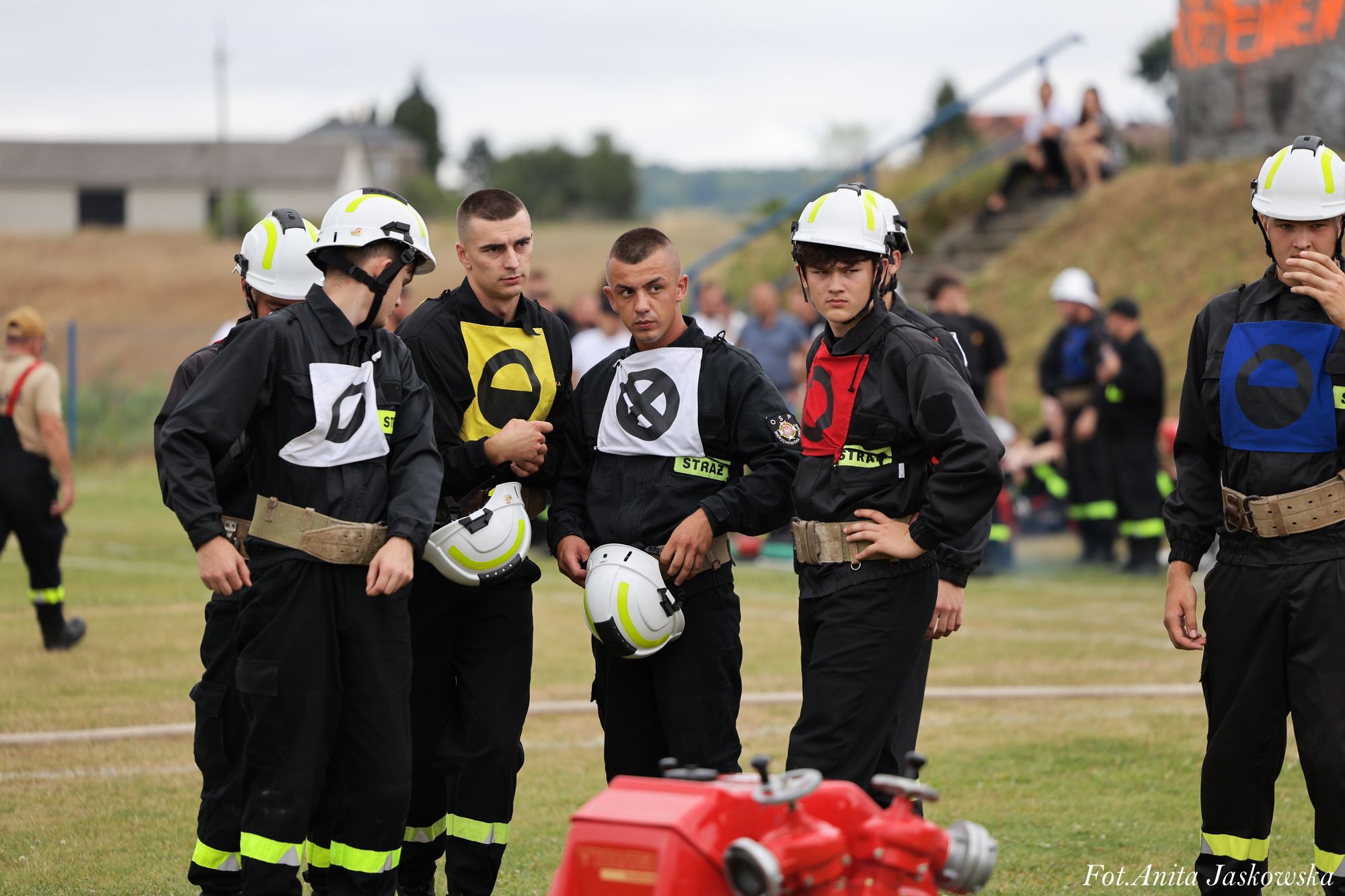 Grupa mężczyzn w czarnych ubraniach strażackich z kaskami, kolorowymi oznaczeniami na piersiach, stojąca na trawiastym polu, w tle rozmyte postacie i budynki.
