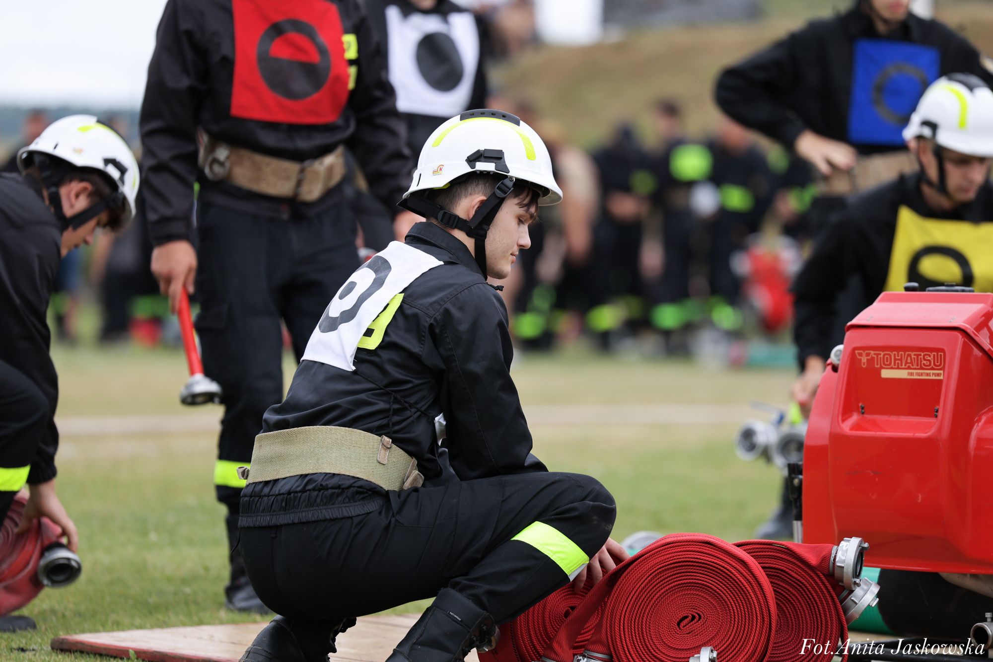 Młodzi strażacy w czarnych mundurach i białych hełmach z kolorowymi oznaczeniami, zwinięte czerwone węże obok czerwonej pompy pożarniczej na trawie.