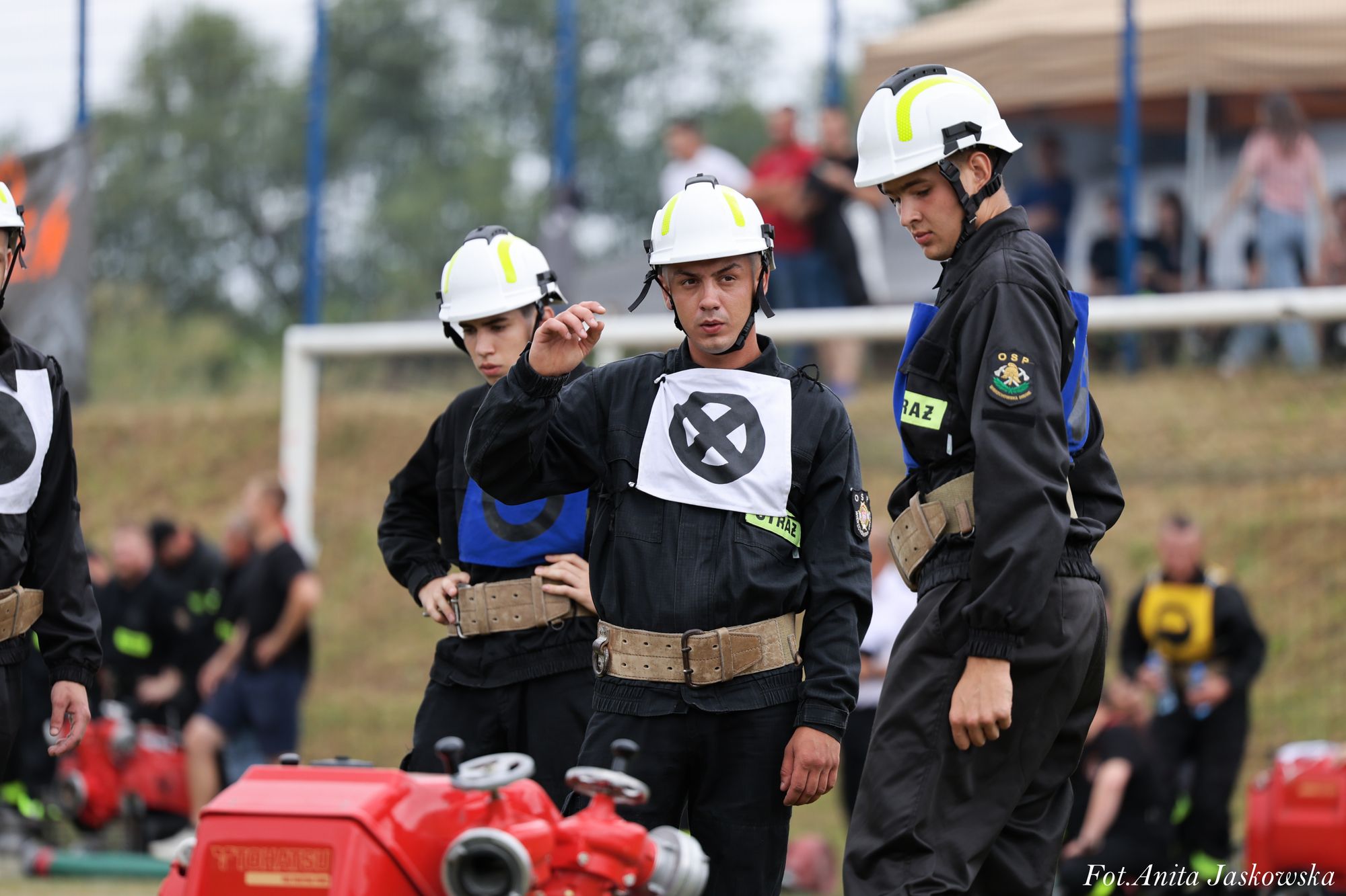 Czterech mężczyzn w czarnych mundurach i białych hełmach, z oznaczeniami OSP i STRAŻ, stoi przy czerwonym sprzęcie na tle rozmytej zieleni i ludzi.