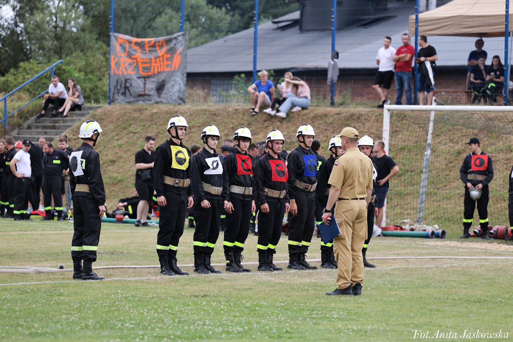 Grupa młodych strażaków w czarnych mundurach, białych hełmach i kolorowych oznaczeniach stoi na trawie, przed nimi mężczyzna w beżowym mundurze, w tle ludzie na trawie i trybunach.