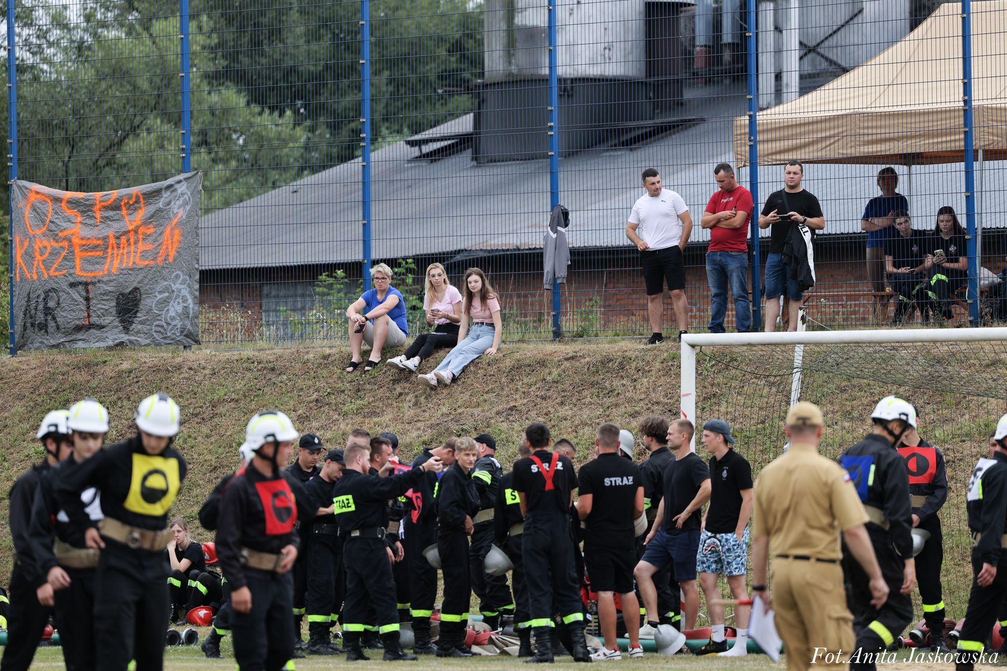 Grupa strażaków w czarnych mundurach z neonowymi elementami i kaskami stoi na murawie przy bramce, na tle ogrodzenia z banerem i siedzącymi oraz stojącymi widzami.