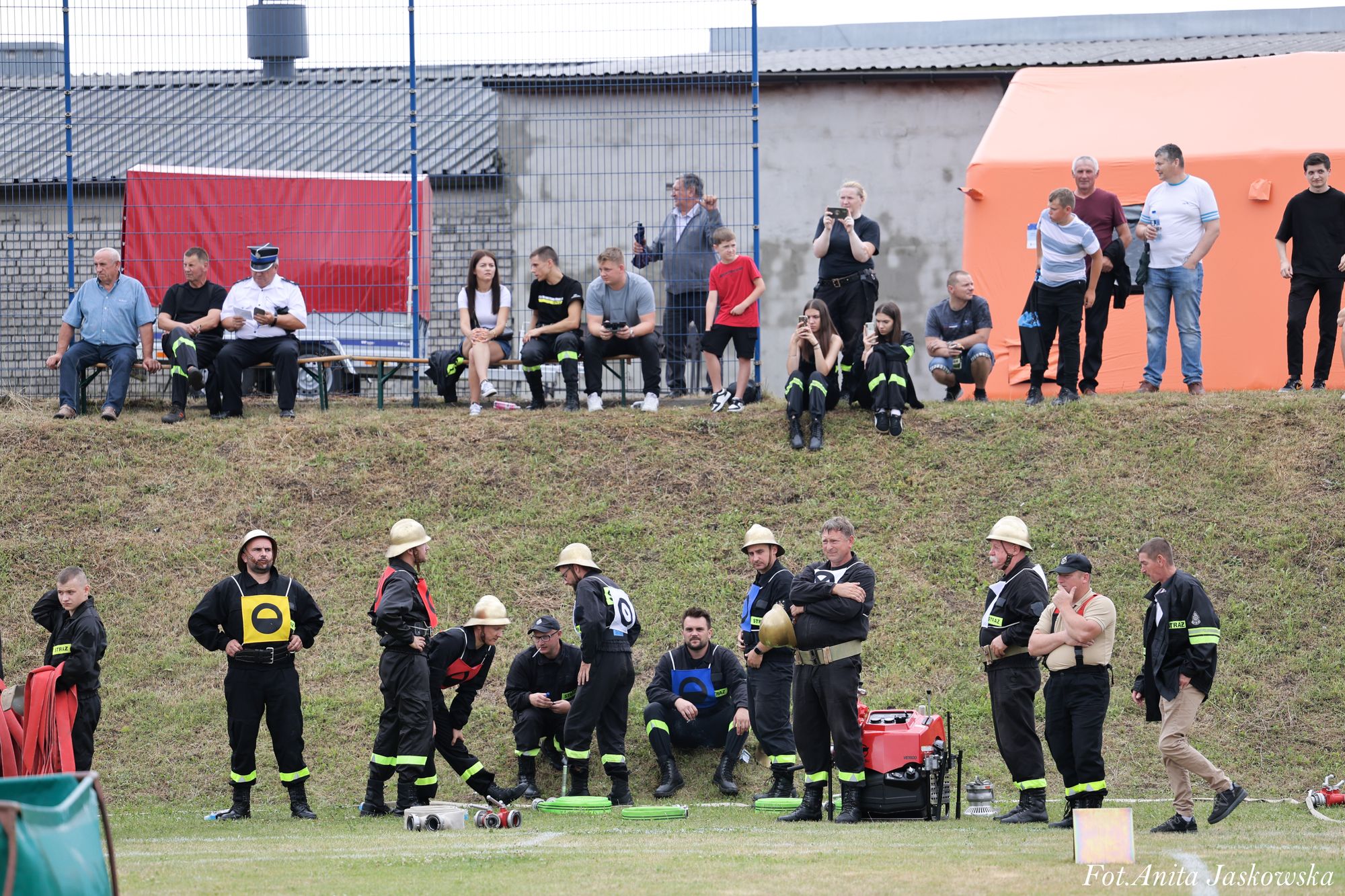 Grupa strażaków w czarnych mundurach z żółtymi i niebieskimi oznaczeniami oraz hełmami stoi i siedzi na trawie, w tle widoczni widzowie i pomarańczowy namiot.