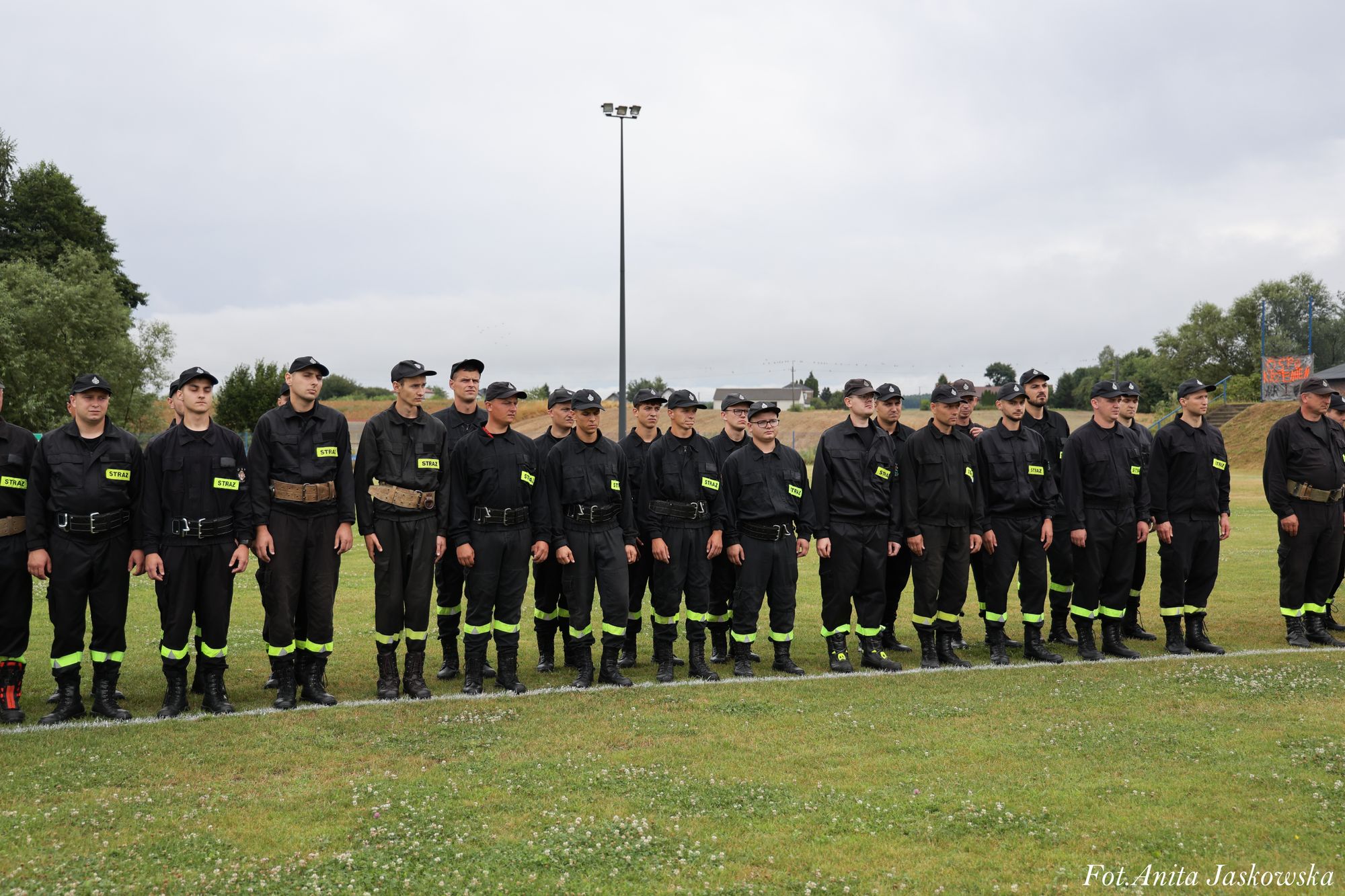 Grupa mężczyzn w czarnych mundurach z odblaskowymi żółtymi oznaczeniami "STRAŻ", stojących na trawie na tle pochmurnego nieba i zielonych drzew.