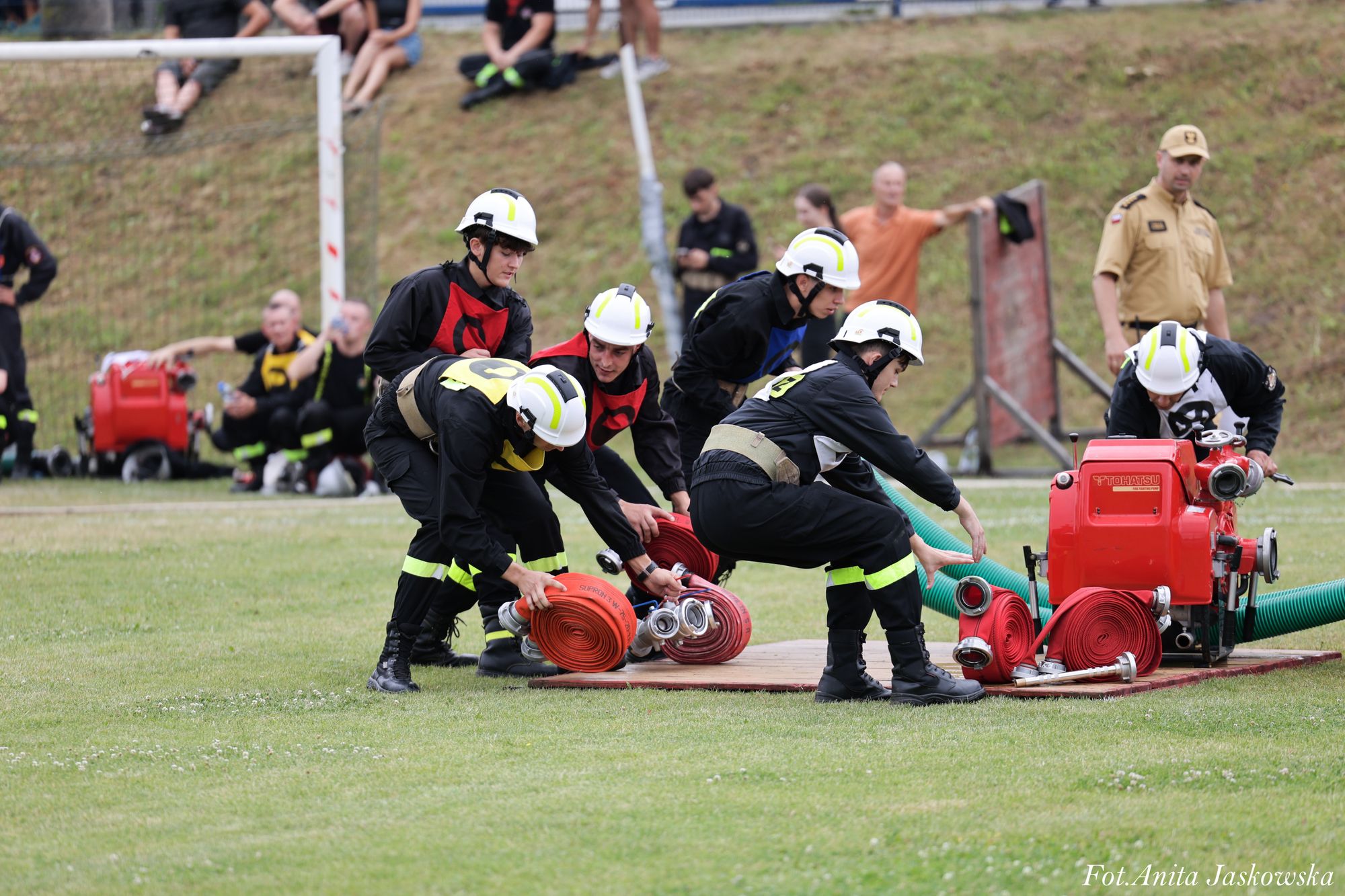 Grupa młodych strażaków w hełmach i ubraniach ochronnych rozwija czerwone zwinięte węże obok czerwonej pompy na trawniku, w tle widoczni obserwujący ludzie i murawa.