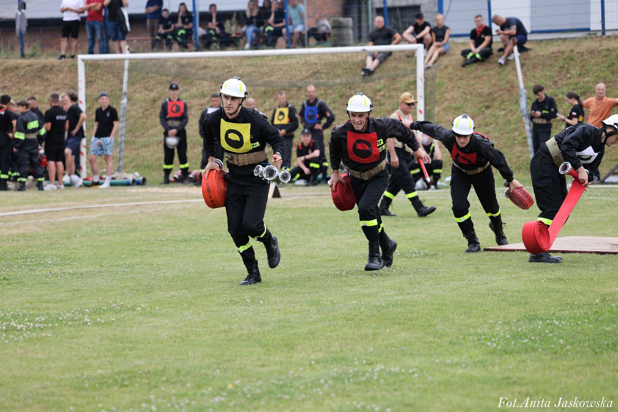Czterech strażaków w kaskach i czarnych mundurach biegnie po trawiastym boisku, każdy trzyma zwinięty czerwony wąż strażacki, w tle ludzie i bramka piłkarska.
