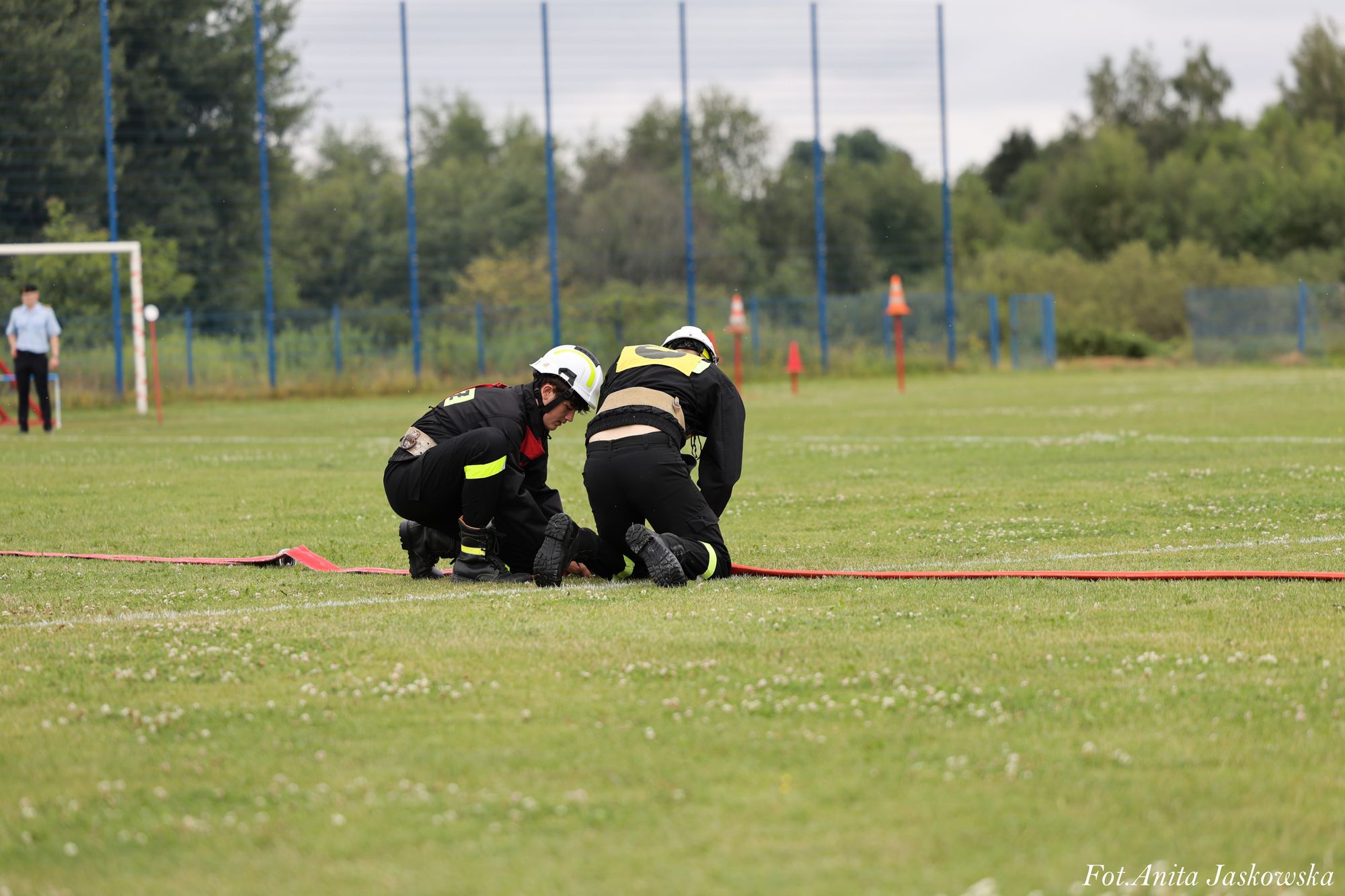 Dwóch mężczyzn w czarnych mundurach i białych hełmach klęczy na zielonej trawie, rozkładając czerwoną linię. W tle bramka i drzewa.
