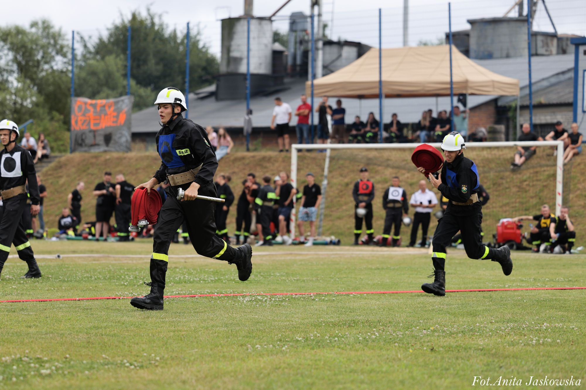Dwóch strażaków w czarnych mundurach i białych hełmach biegnie po zielonym boisku, każdy niesie zwinięty czerwony wąż strażacki, w tle stoją kibice i budynki.