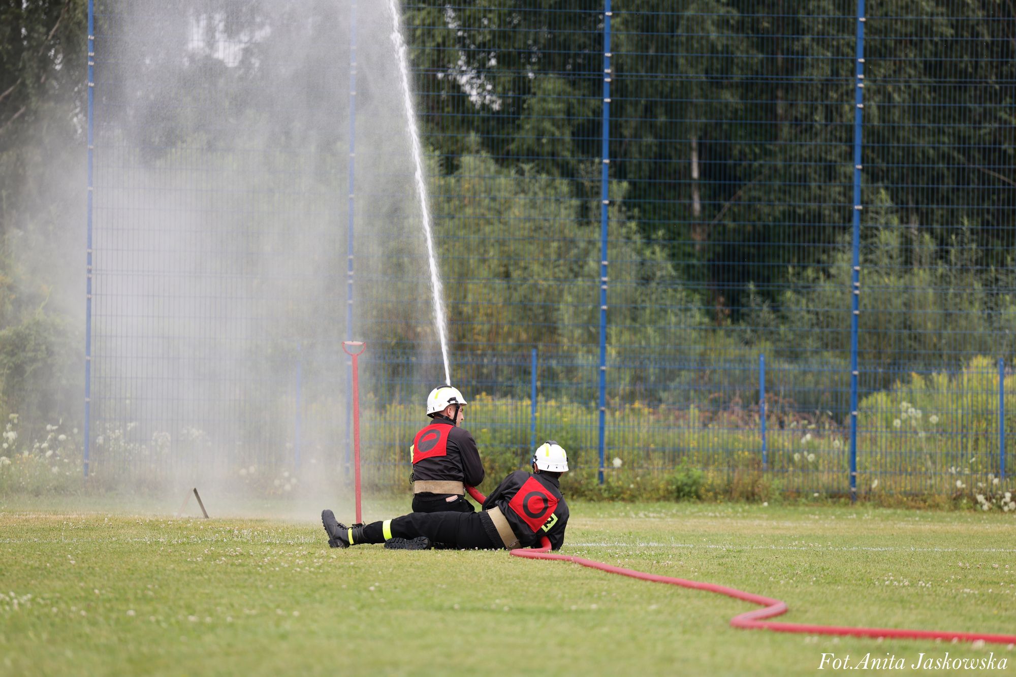 Dwóch strażaków w czarnych mundurach i białych hełmach obsługuje czerwoną linię wodną na zielonym trawiastym boisku przy ogrodzeniu z siatki.