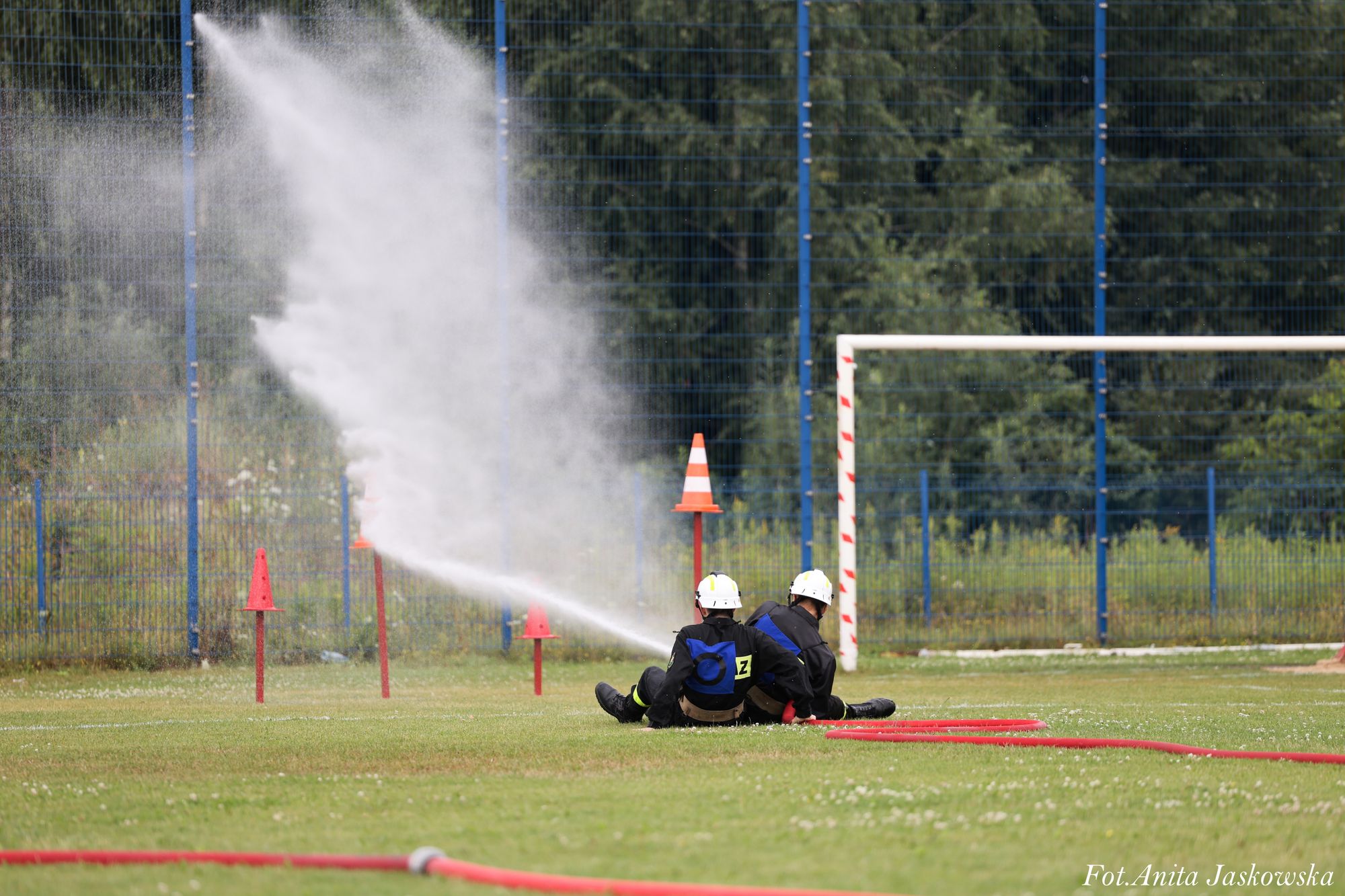 Dwóch strażaków w czarnych uniformach i białych hełmach siedzi na trawie, trzymając strumień wody z węża strażackiego, obok słupki i bramka na boisku.