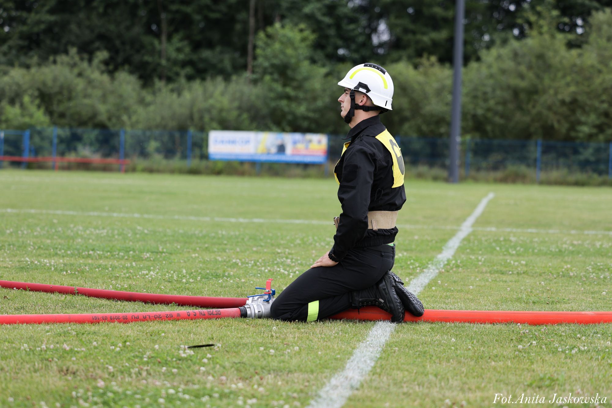 Mężczyzna w czarnym uniformie i białym kasku klęczy na zielonej trawie obok czerwonych węży strażackich, w tle zieleń i ogrodzenie.
