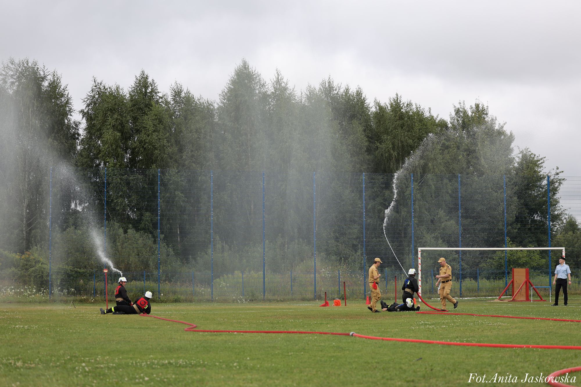 Strażacy w kaskach i mundurach ćwiczą gaszenie wodą na zielonym boisku z bramką, w tle wysoki płot i drzewa pod pochmurnym niebem.