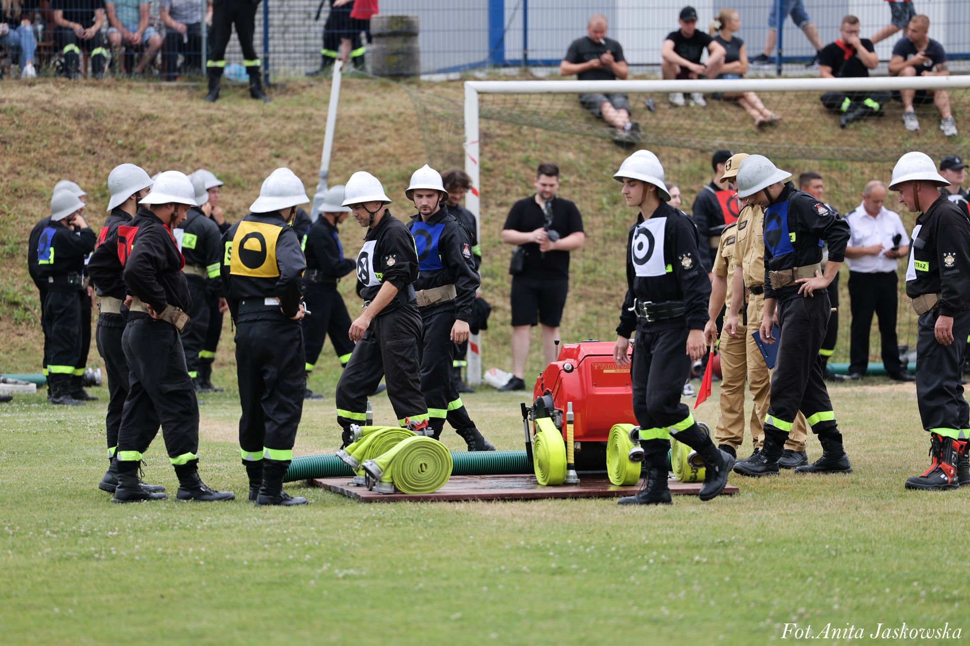 Grupa osób w czarnych mundurach z białymi hełmami i kolorowymi oznaczeniami stoi na trawie wokół złożonych zielonych węży strażackich i czerwonego sprzętu.