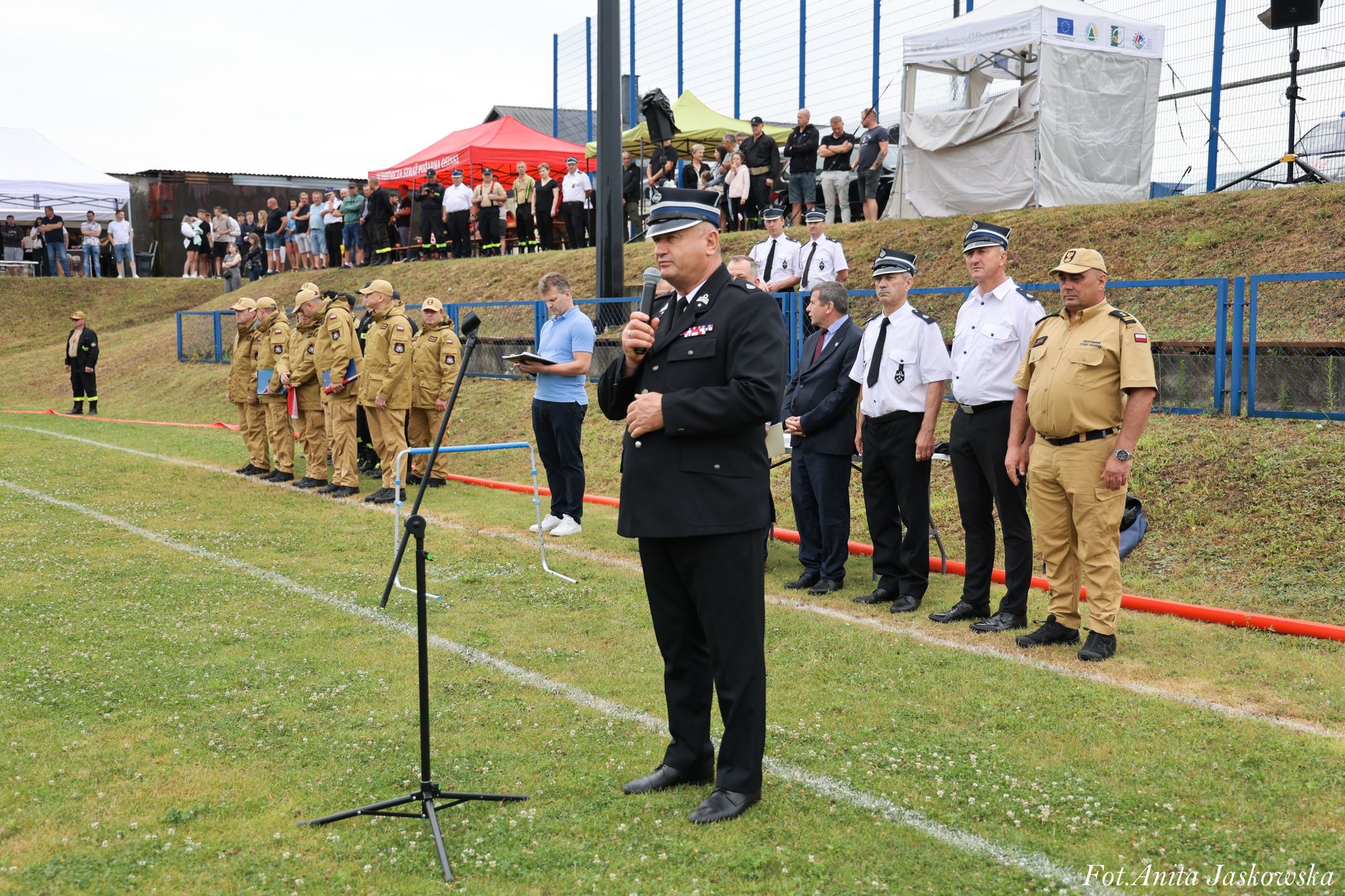Mężczyzna w czarnym mundurze z mikrofonem stoi na trawie, za nim szeregi mężczyzn w mundurach beżowych i białych oraz grupa ludzi na tle ogrodzenia i namiotów.