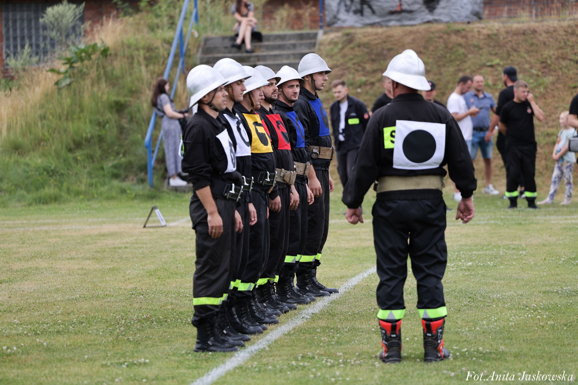 Siedmiu mężczyzn w czarnych mundurach i białych hełmach stoi w szeregu na zielonej trawie, jeden mężczyzna stoi przodem do nich, na mundurach kolorowe oznaczenia.