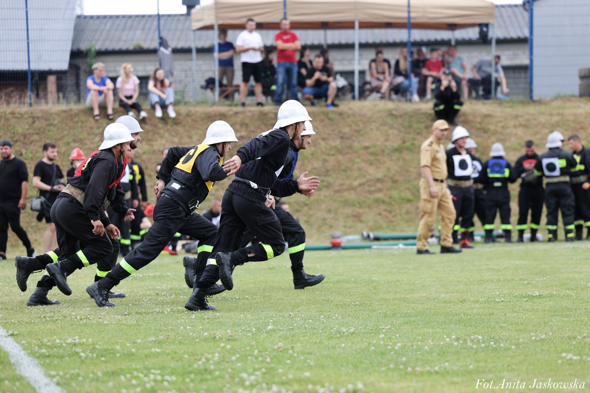Grupa mężczyzn w czarnych mundurach i białych hełmach biegnie po trawie, w tle ludzie siedzą i stoją na pagórku.