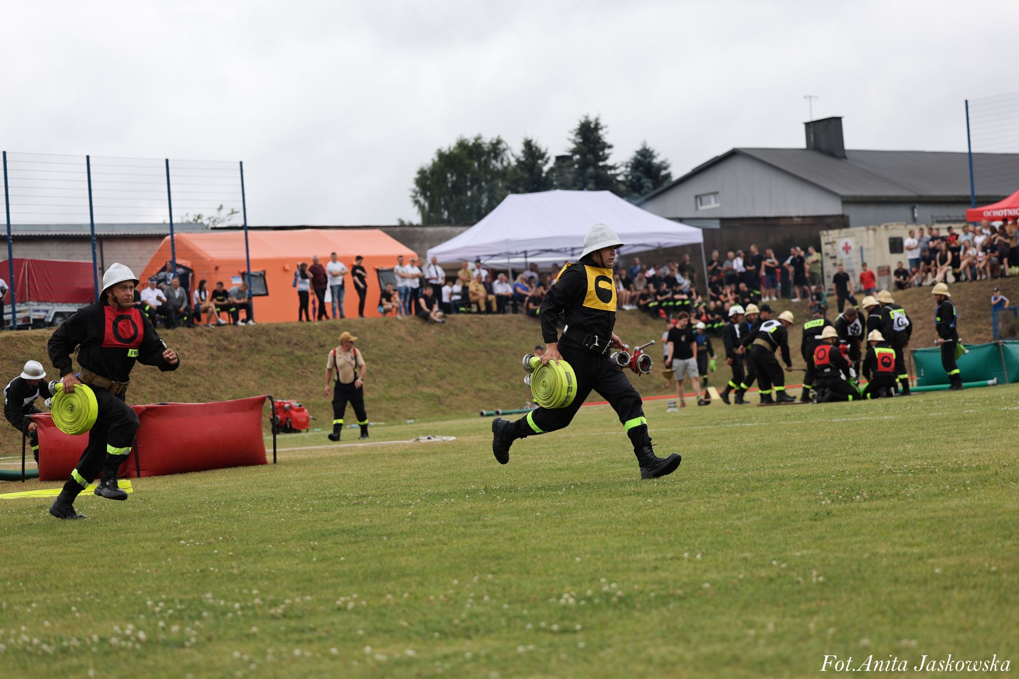 Dwóch strażaków w hełmach i czarnych mundurach z kolorowymi kamizelkami biegnie z żółtymi zwiniętymi węzłami, na tle zielonej trawy i widowni pod namiotem.