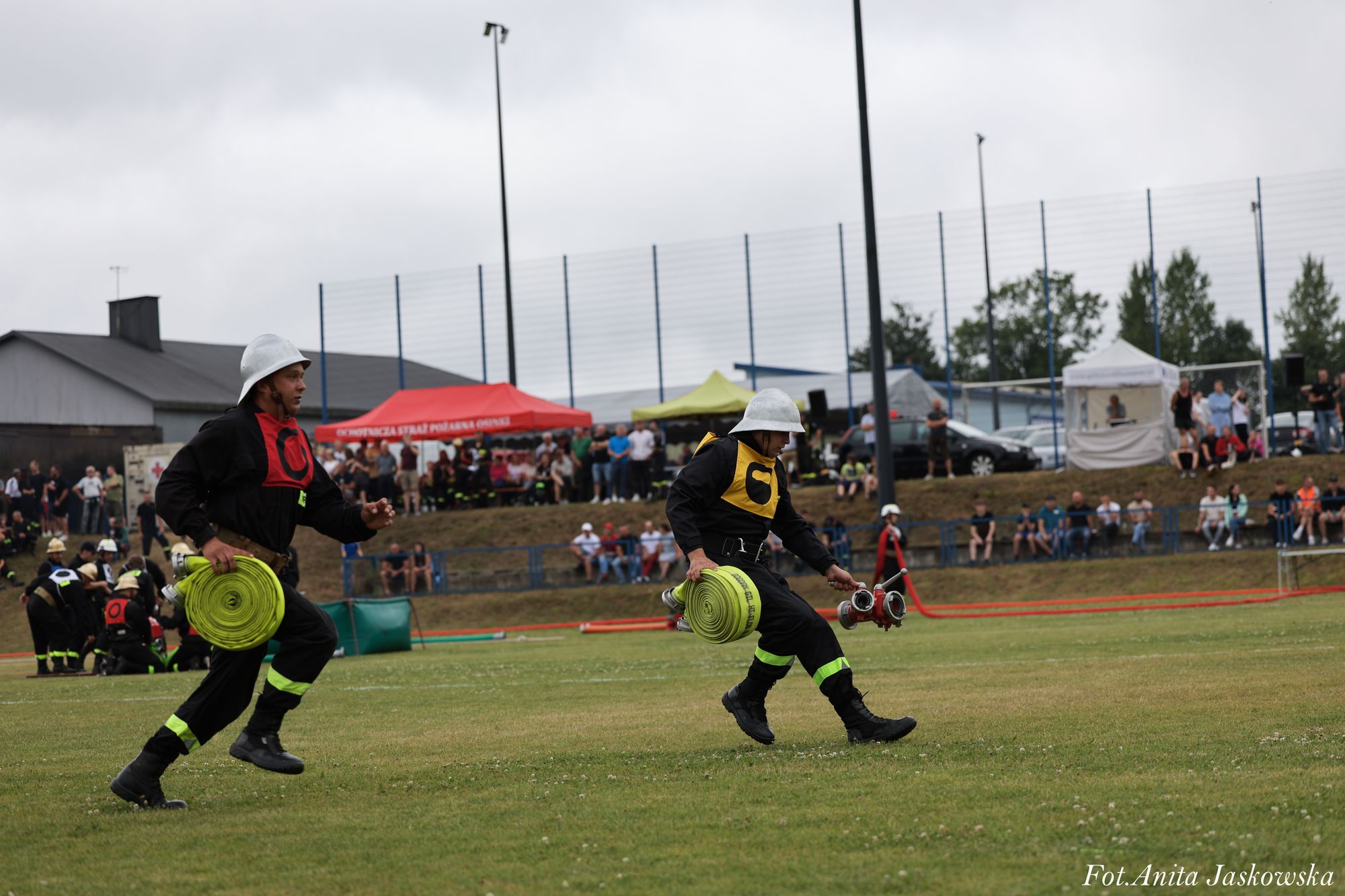 Dwóch strażaków w kaskach i ciemnych mundurach biegnie po zielonym boisku, niosąc żółte zwinięte węże strażackie, w tle widoczna grupa ludzi i namioty.