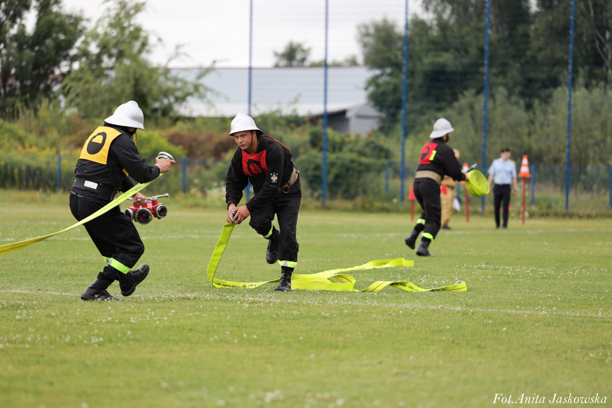 Trzech mężczyzn w czarnych mundurach i białych hełmach rozwija żółte węże strażackie na zielonym boisku, w tle drzewa i ogrodzenie.