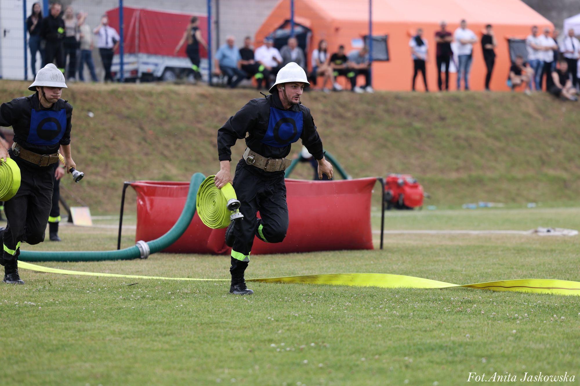 Dwóch mężczyzn w hełmach i czarnych strojach z niebieskimi oznaczeniami, biegnących z żółtymi zwiniętymi wężami strażackimi na zielonym tle trawnika.