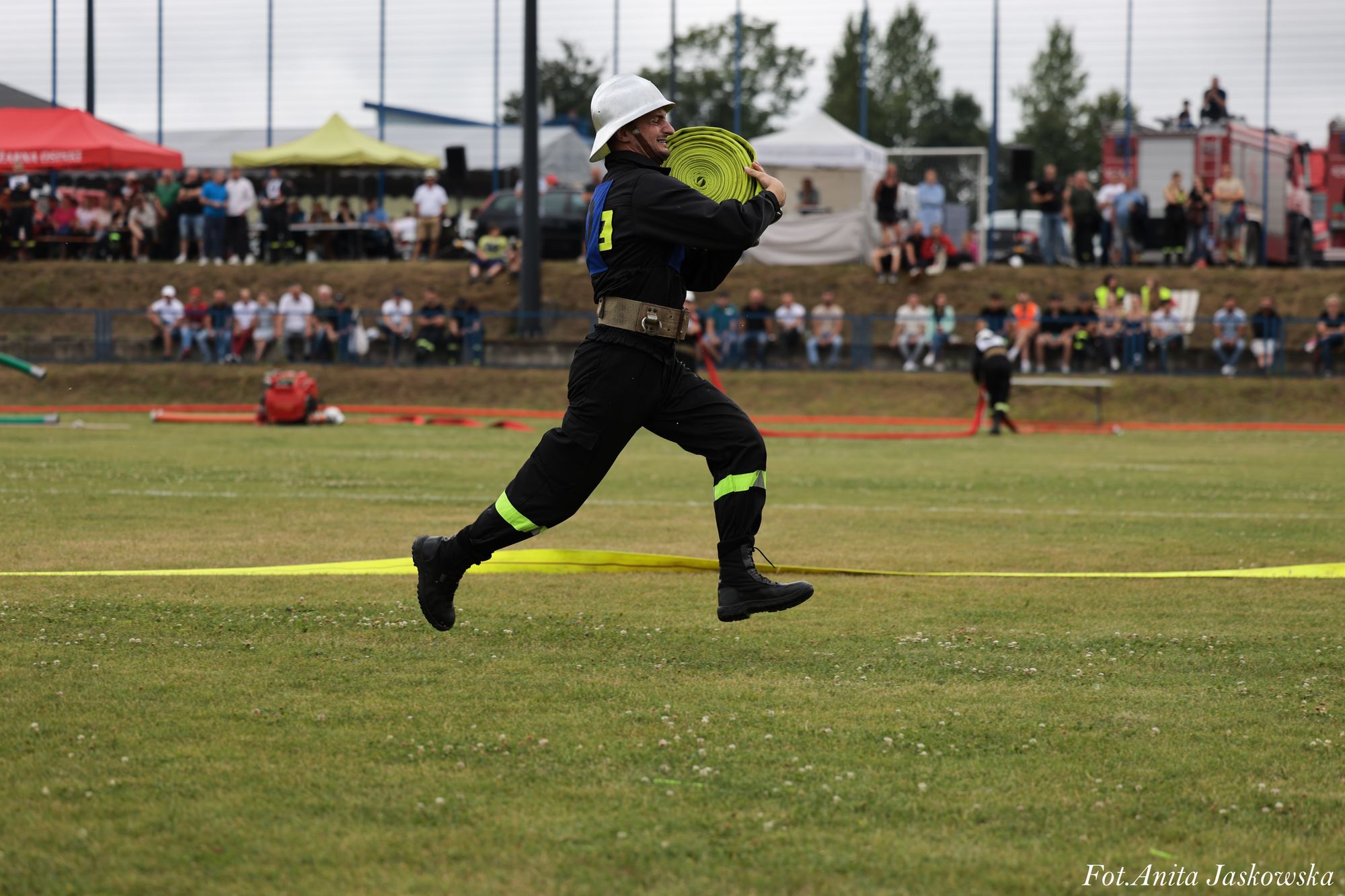 Mężczyzna w czarnym kombinezonie i białym hełmie biegnie po zielonym boisku, trzymając zwinięty żółty wąż strażacki, w tle widoczna publiczność i namioty.