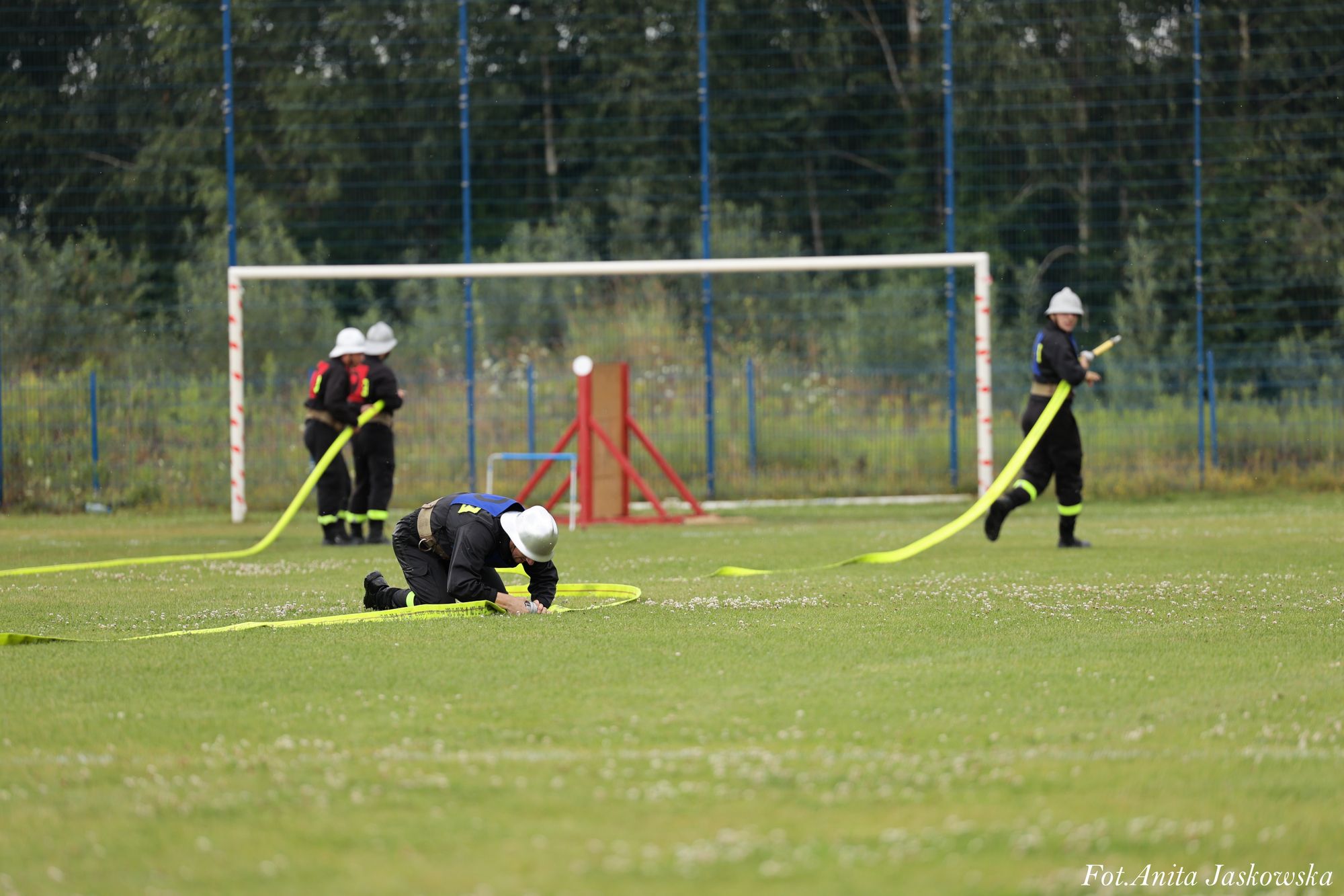 Cztery osoby w czarnych mundurach i białych hełmach na zielonym boisku, jedna klęczy, pozostałe trzymają żółte węże strażackie, w tle bramka i siatka.