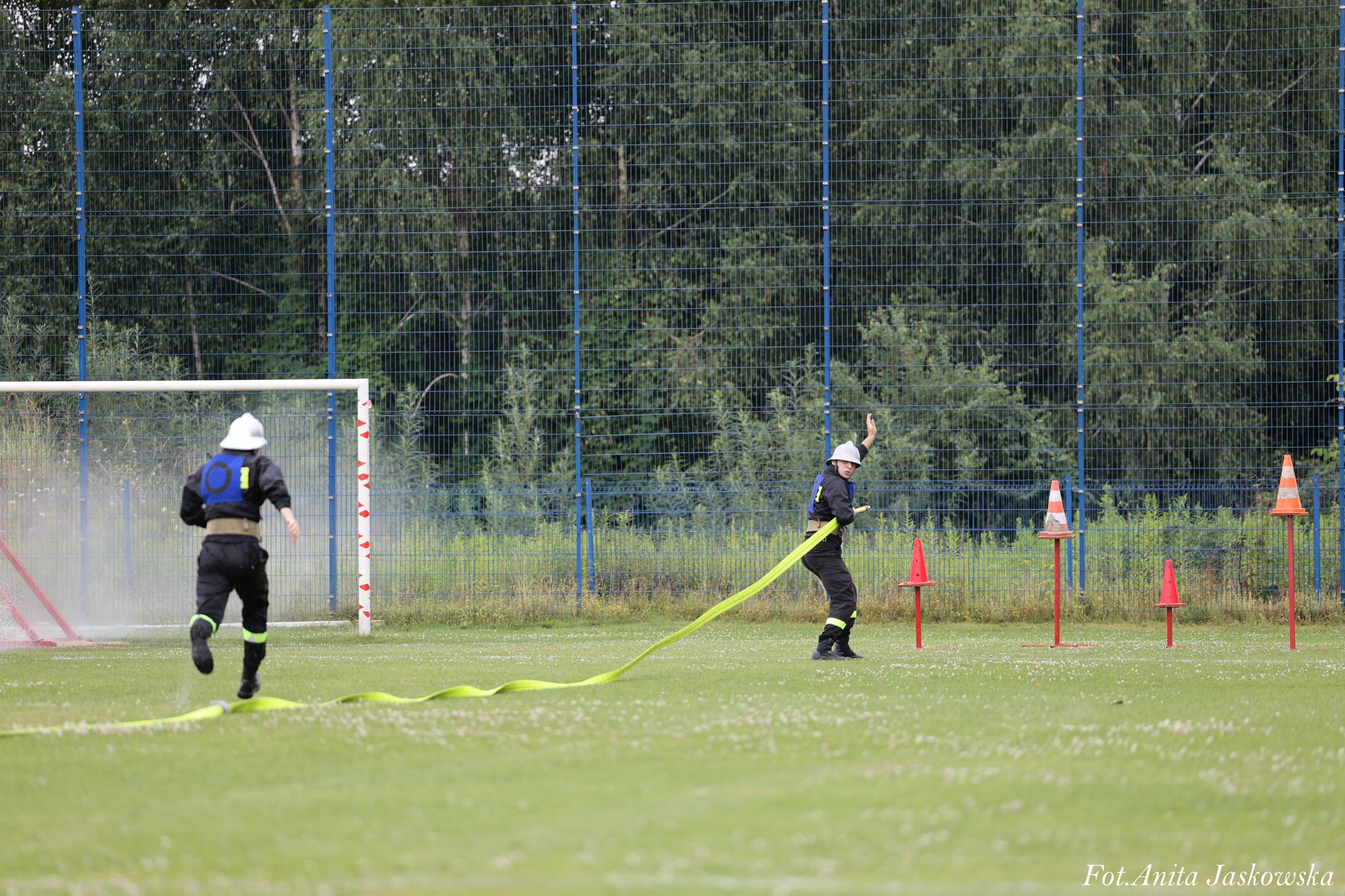 Dwóch strażaków w czarnych mundurach i białych hełmach na zielonym boisku, jeden biegnie, drugi trzyma żółty wąż strażacki, w tle metalowe bramki i zielone drzewa.