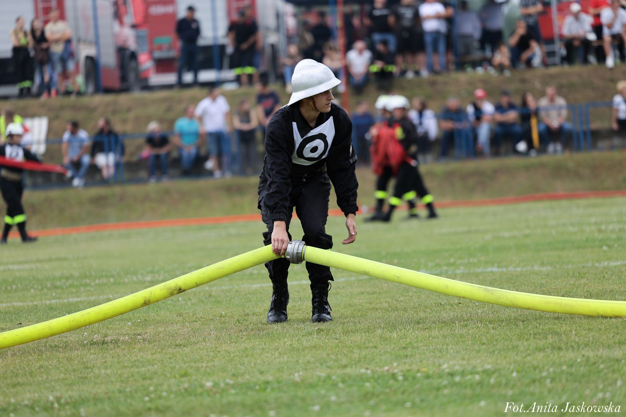 Mężczyzna w czarnym ubraniu i białym hełmie łączy dwa żółte węże strażackie na zielonej trawie, w tle rozmyci ludzie i wozy strażackie.