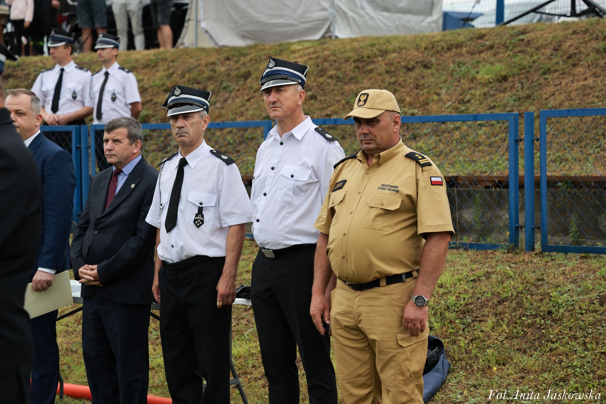 Czterech mężczyzn, dwóch w mundurach białych z czarnymi krawatami i rogatywkami, jeden w beżowym mundurze z czapką, dwóch w garniturach, na tle trawy i niebieskiego ogrodzenia.