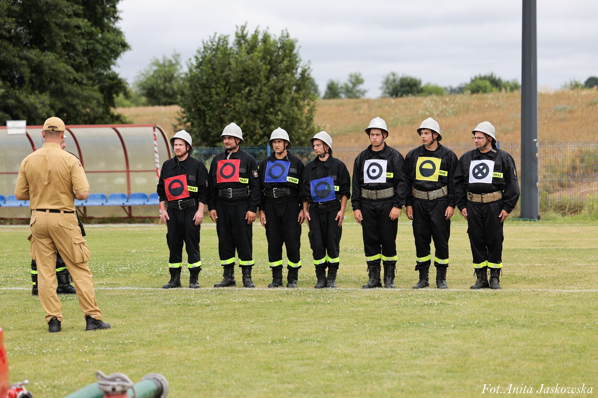 Siedmiu mężczyzn w czarnych mundurach i białych hełmach stoi na trawie w jednej linii, przed nimi stoi mężczyzna w beżowym uniformie, w tle drzewa i niebo.