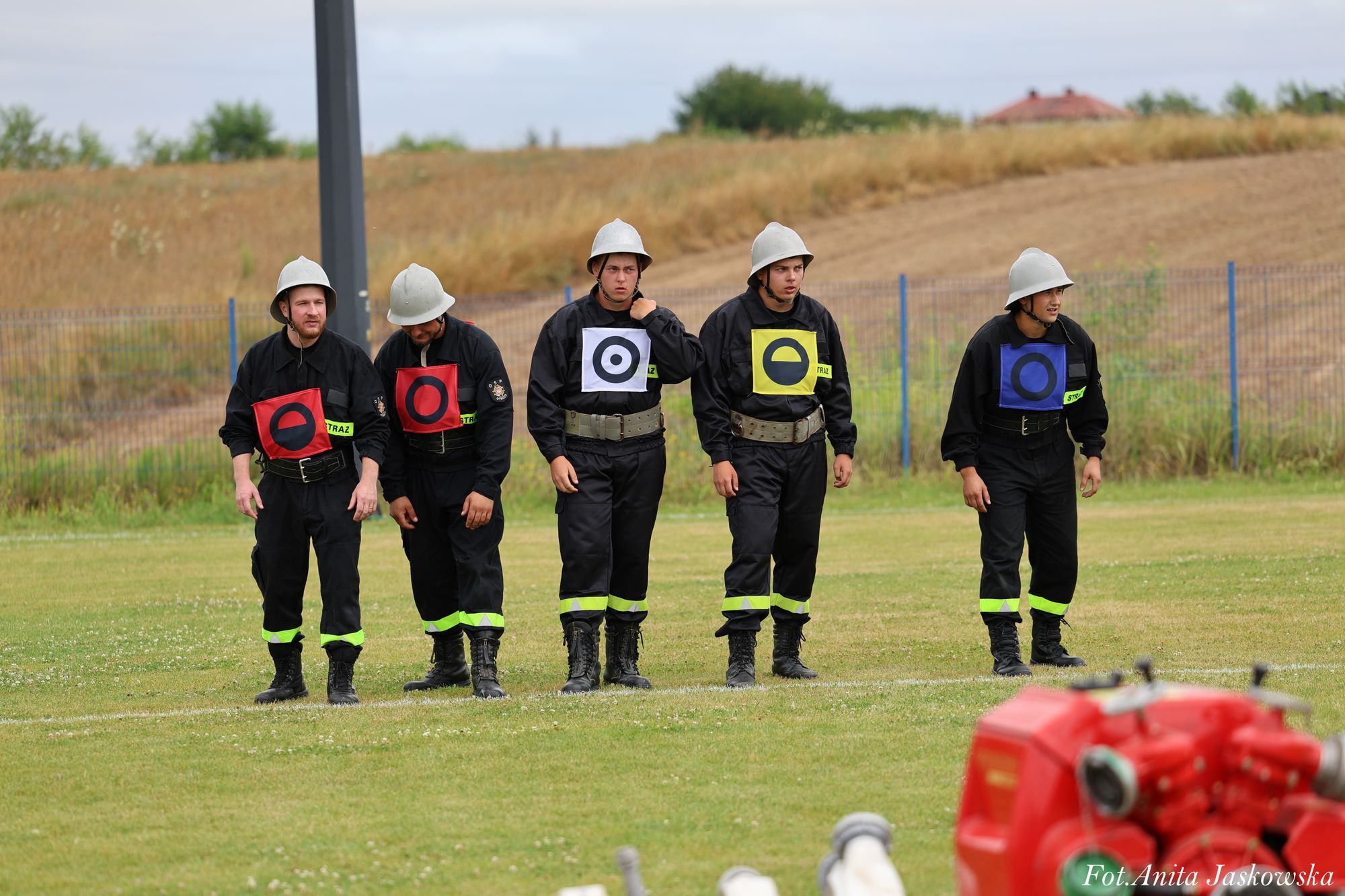 Pięciu mężczyzn w czarnych mundurach strażackich z kolorowymi oznaczeniami i hełmami stoi na zielonym polu z rozmytym czerwonym sprzętem w pierwszym planie.