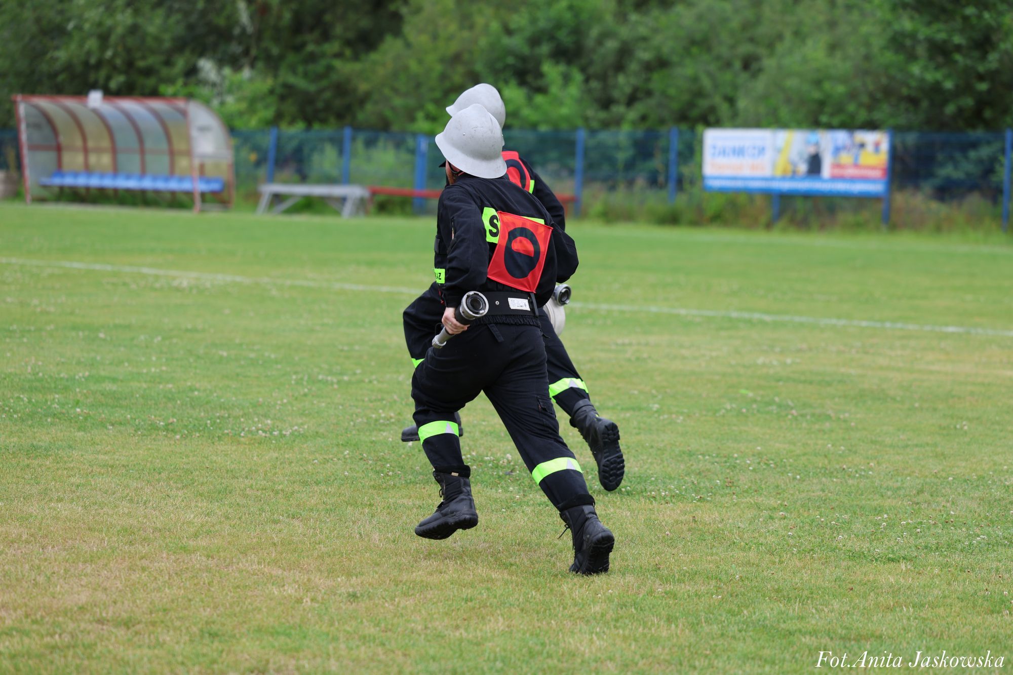 Dwaj strażacy w czarnych mundurach z odblaskowymi pasami i białych hełmach biegną po zielonym trawniku, każdy trzyma metalową rurę. W tle niebieski płot i zadaszona ławka.