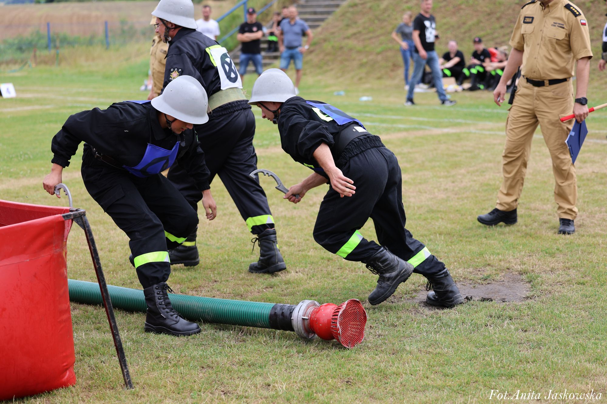 Trzej mężczyźni w czarnych mundurach z odblaskowymi paskami i szarych hełmach przy zielonym wężu strażackim na trawie, po prawej stojący mężczyzna w beżowym mundurze.