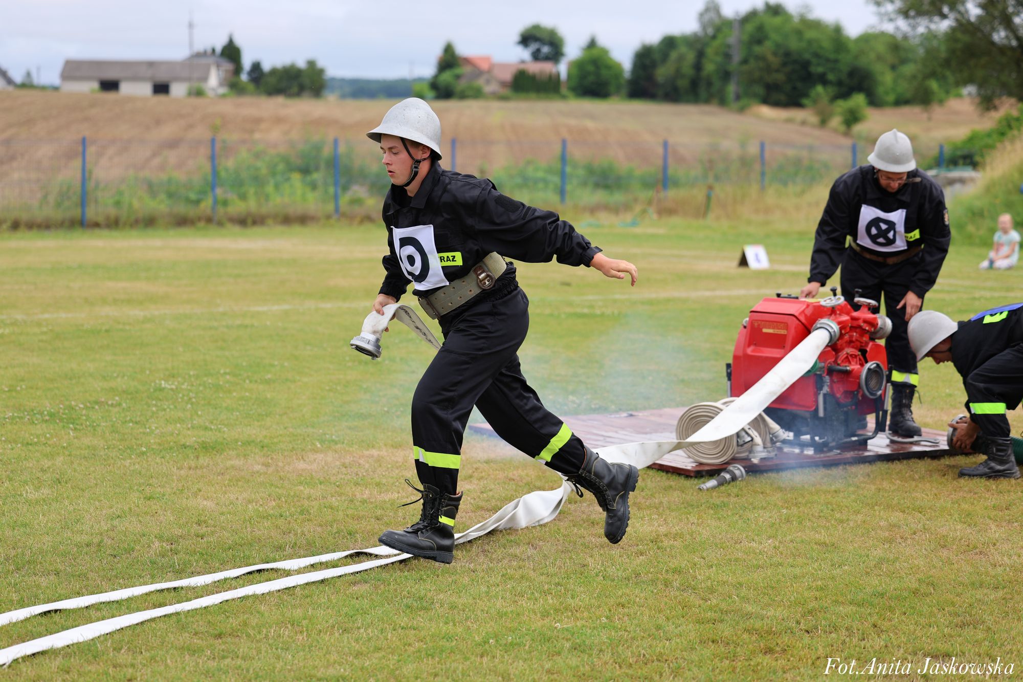 Mężczyzna w czarnym ubraniu strażackim i hełmie biegnie po trawie z białym wężem strażackim, w tle dwaj inni mężczyźni przy czerwonej pompie wodnej na tle pola i ogrodzenia.