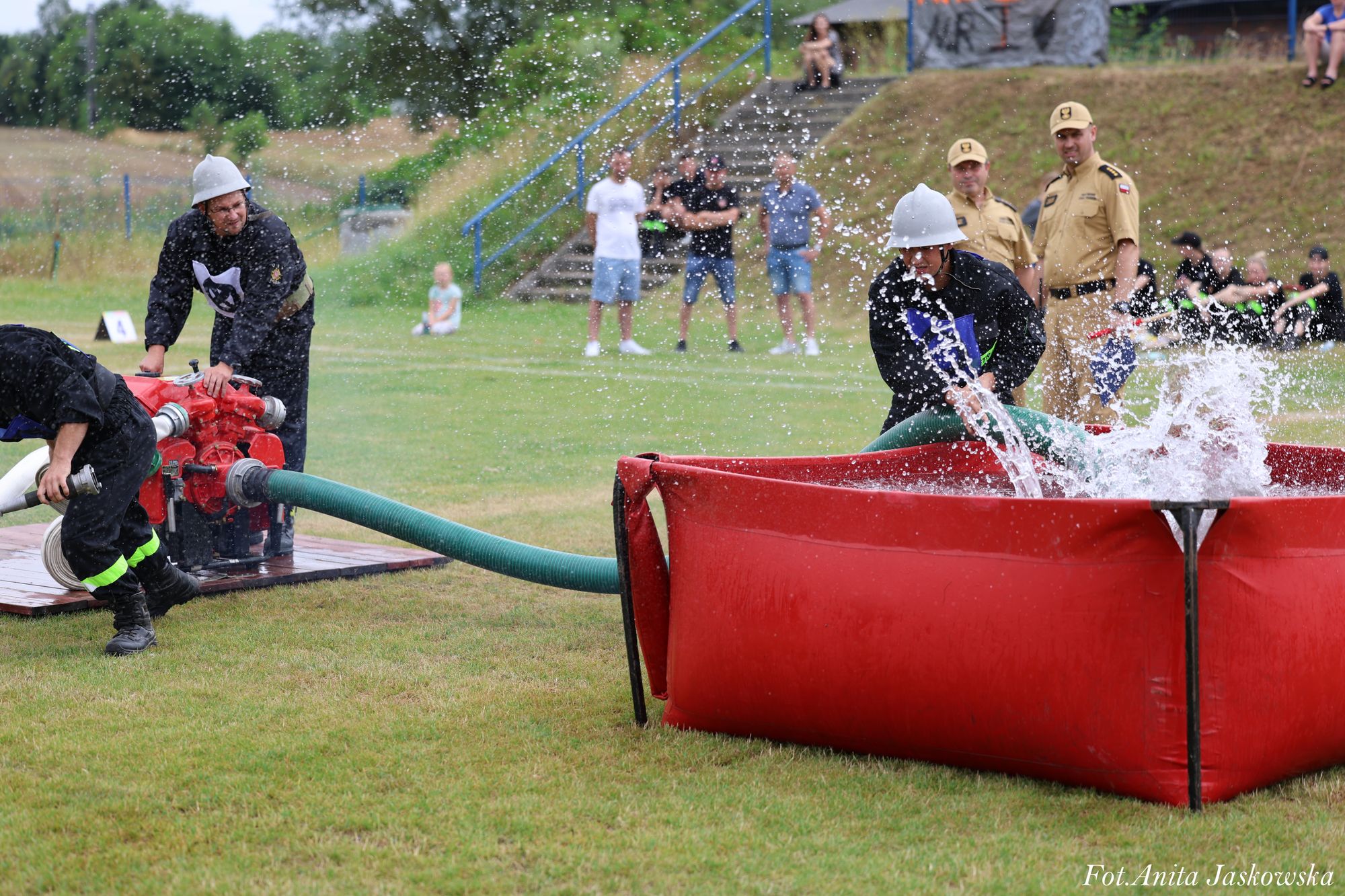 Strażacy w czarnych mundurach i hełmach przy czerwonym zbiorniku z wodą na zielonej trawie, w tle ludzie na schodach i stojący mężczyźni w beżowych mundurach.