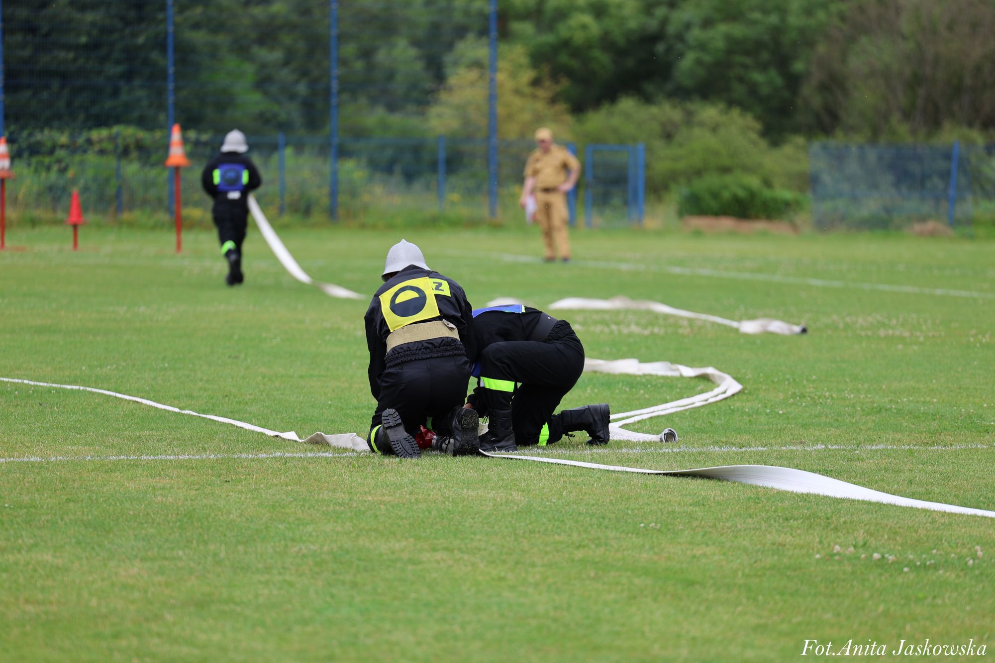 Dwie osoby w czarnych ubraniach ochronnych klęczą na zielonej trawie przy białej linii, rozkładając długi biały wąż strażacki, w tle druga osoba idzie przed siebie, a trzecia stoi.