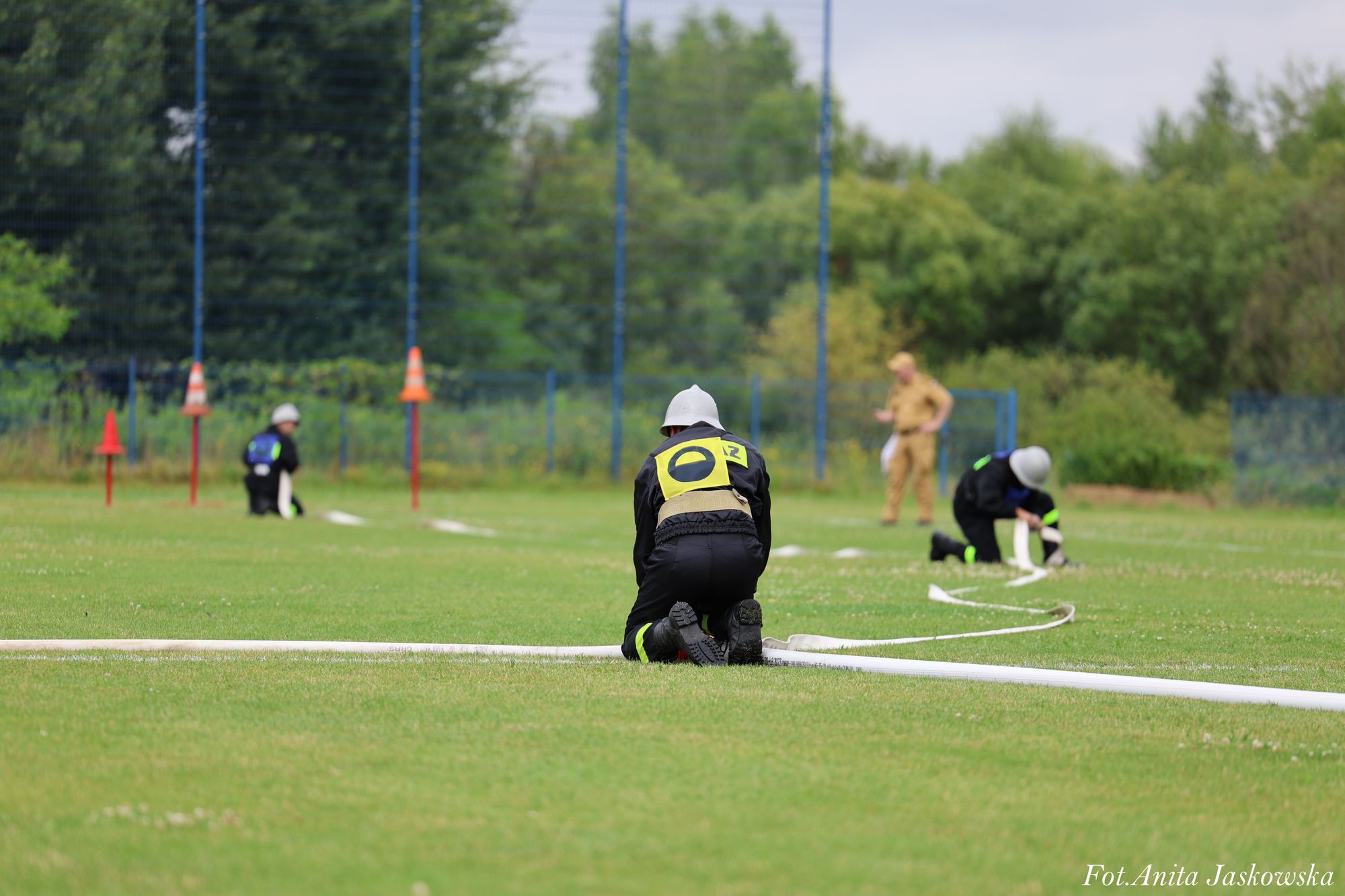 Trzy osoby w kaskach i ubraniach ochronnych klęczą na trawie rozkładając długie białe węże, w tle drzewa i ogrodzenie z niebieskimi słupkami.