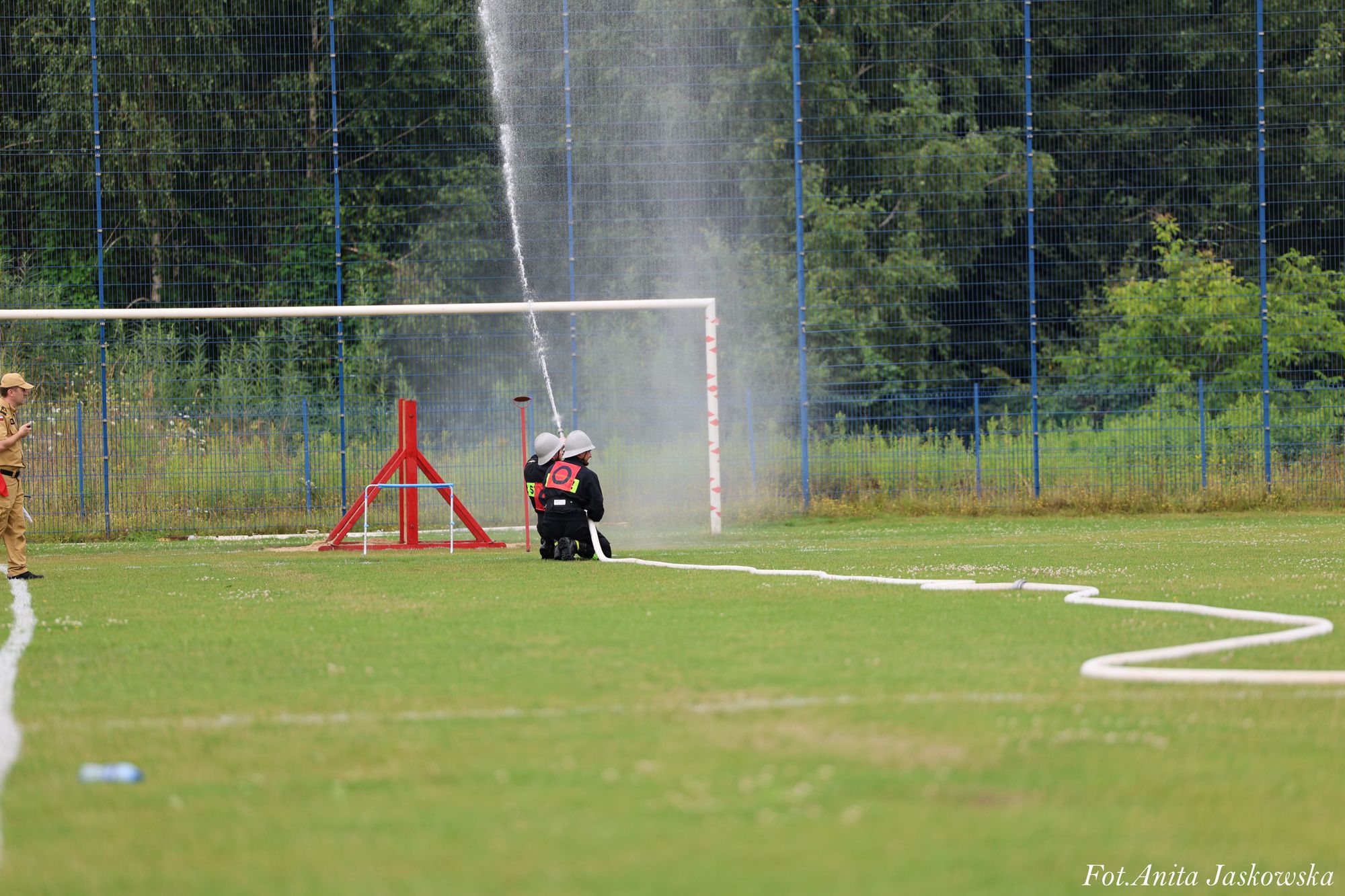Dwoje osób w czarnych strojach i białych hełmach obsługuje biały prąd wody z węża na zielonym boisku, obok stoi mężczyzna w brązowym ubraniu, w tle niebieska siatka i drzewa.