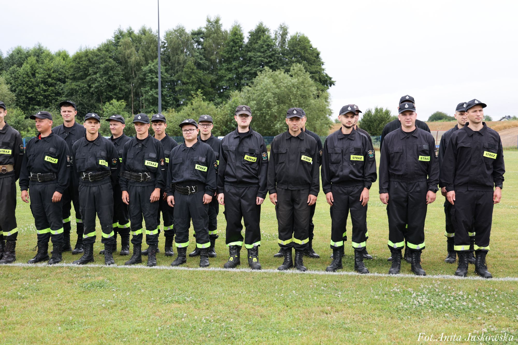 Grupa mężczyzn w czarnych mundurach z żółtymi napisami "STRAŻ", stojących na trawie na tle zielonych drzew i nieba.
