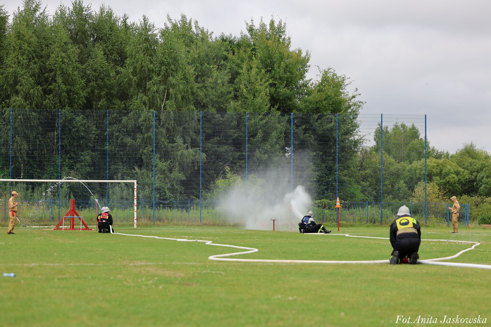 Strażacy w czarnych mundurach z żółtymi oznaczeniami na kolanach na zielonym boisku trawiastym, z mgłą wody, metalowym ogrodzeniem i drzewami w tle.
