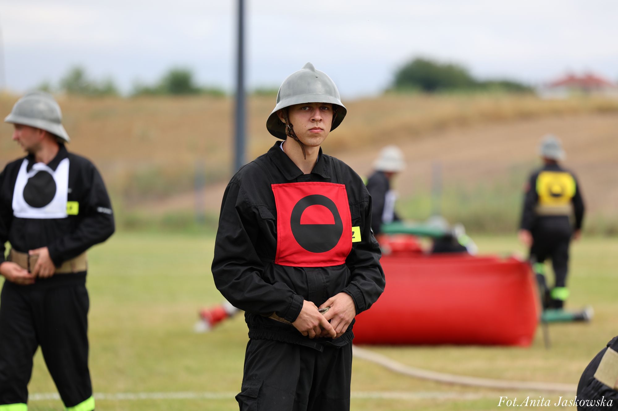 Młody mężczyzna w czarnym mundurze, szarym hełmie i czerwonej naramiennik z czarnym symbolem, stoi na trawie, w tle inni w podobnych strojach i czerwony zbiornik.