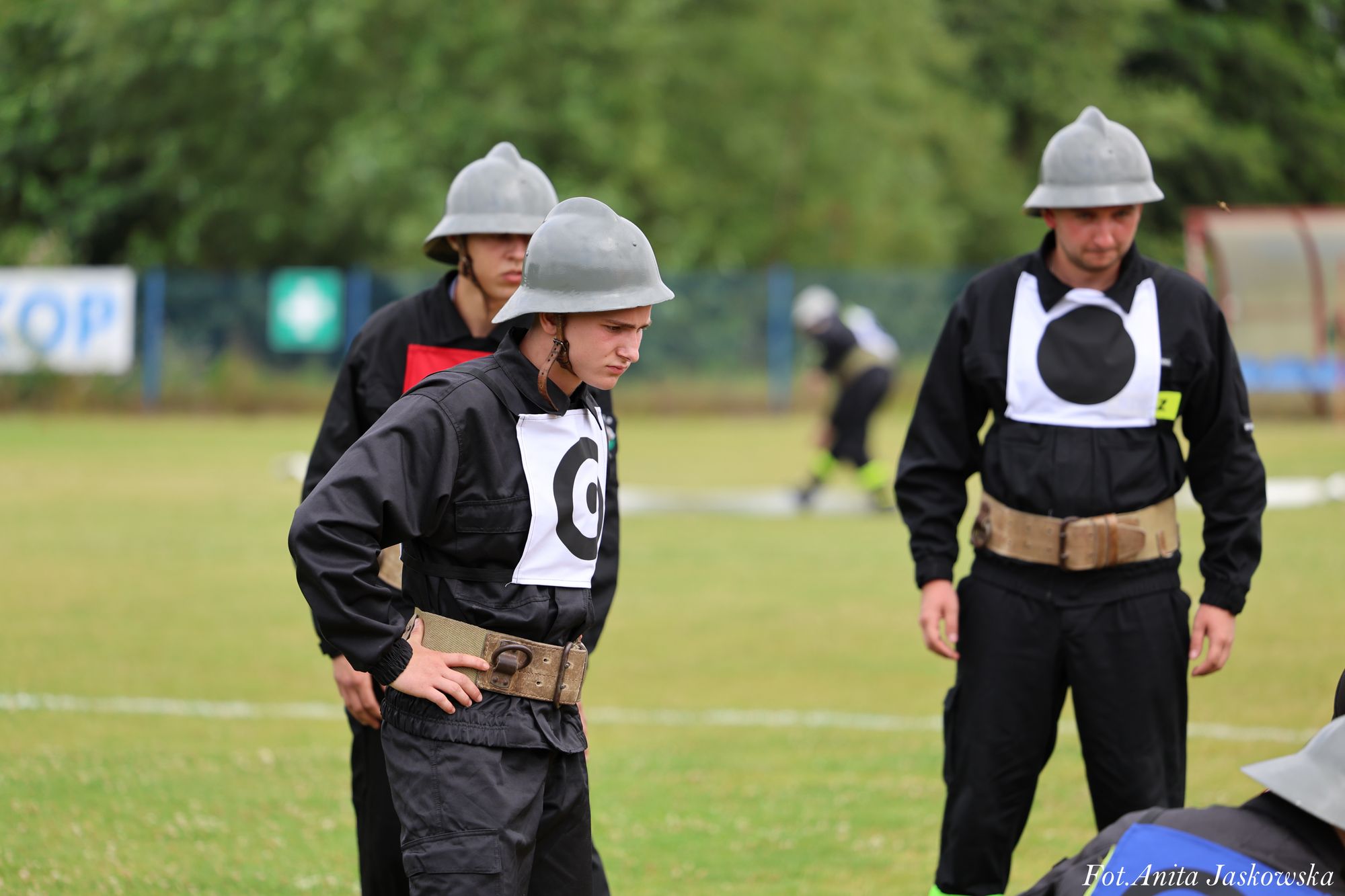 Trzech mężczyzn w czarnych mundurach, szarych hełmach i z oznaczeniami na piersiach stoi na zielonym boisku, w tle rozmyta postać w mundurze.