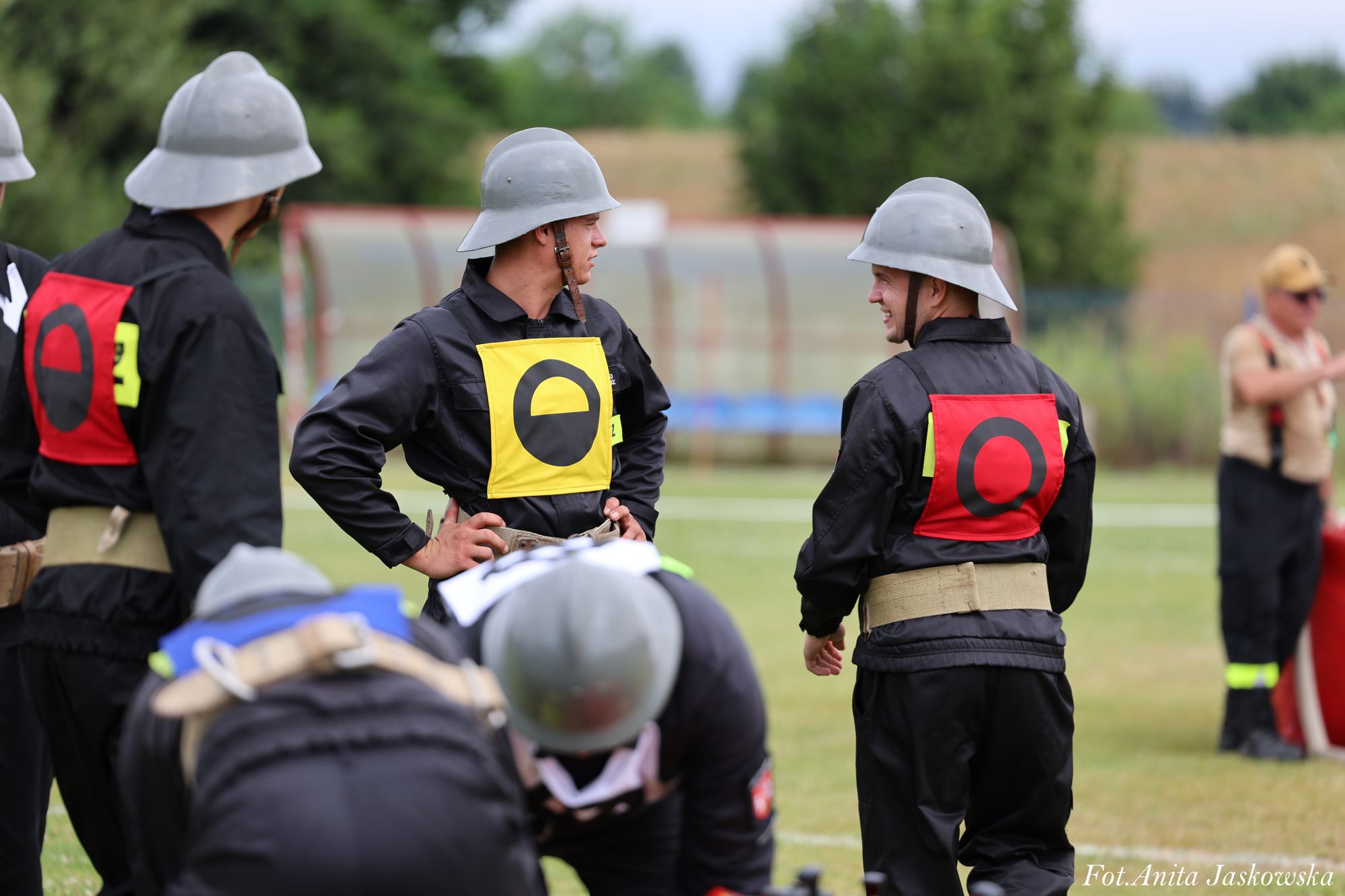 Grupa mężczyzn w czarnych mundurach i szarych hełmach, z kolorowymi oznaczeniami na plecach, stojących na trawiastym boisku na tle zielonych drzew.
