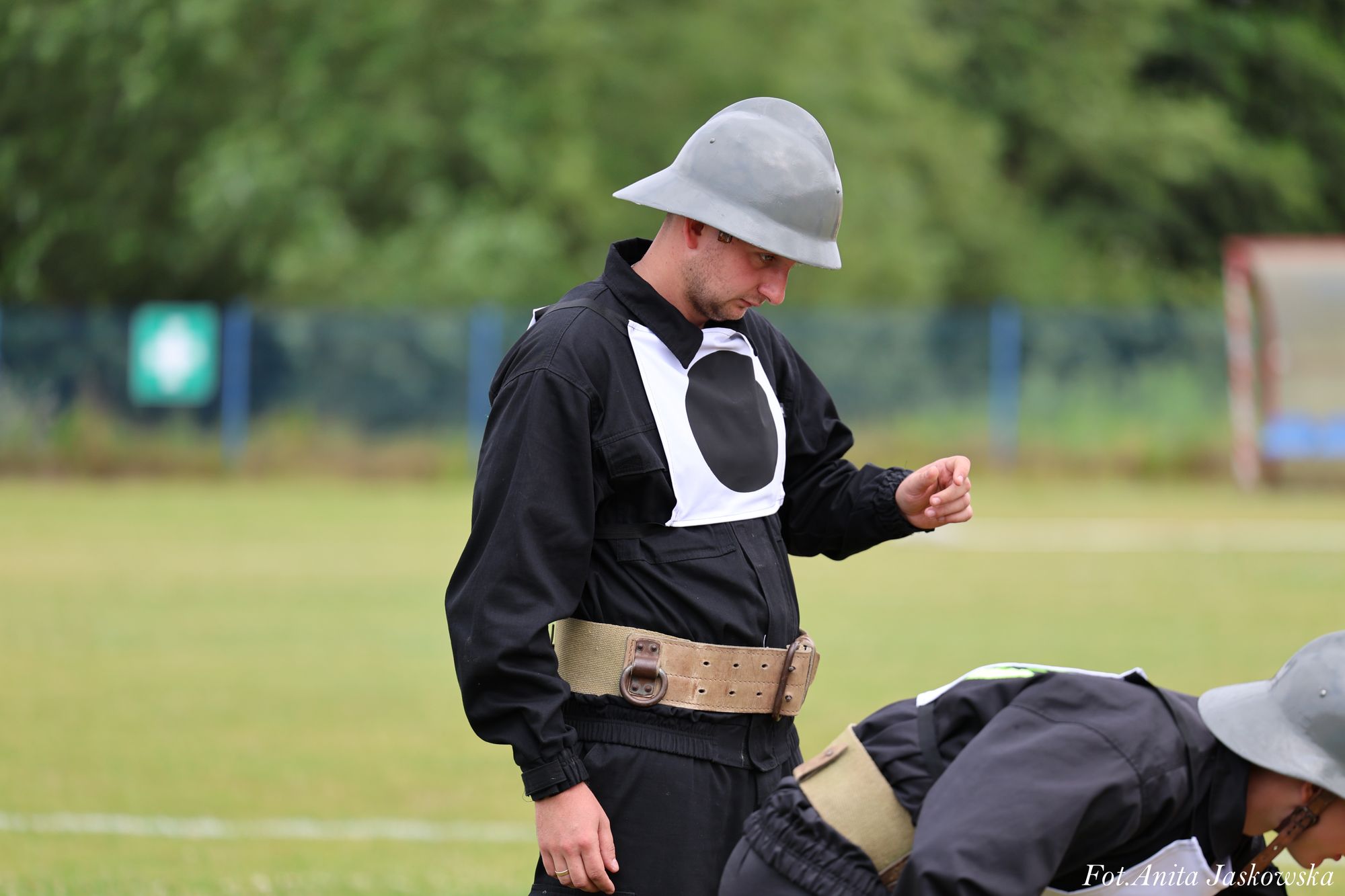 Mężczyzna w czarnym mundurze z szerokim beżowym pasem i szarym hełmem, z białym oznaczeniem na piersi, stoi na zielonym trawiastym boisku, obok pochylona osoba.