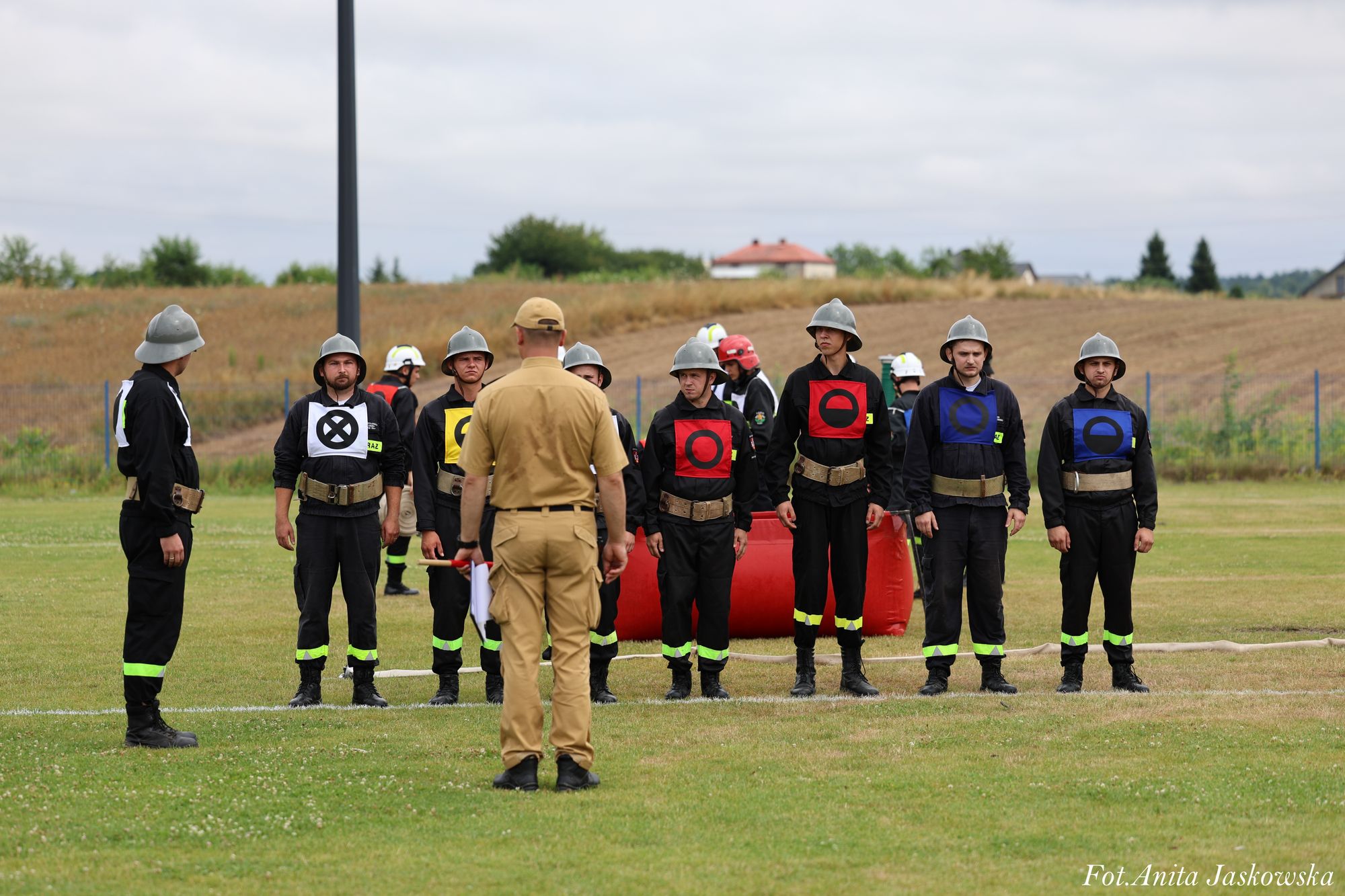 Grupa mężczyzn w czarnych mundurach i hełmach stoi na trawie, obok nich stoi mężczyzna w jasnobrązowym mundurze, na tle pola i nieba.