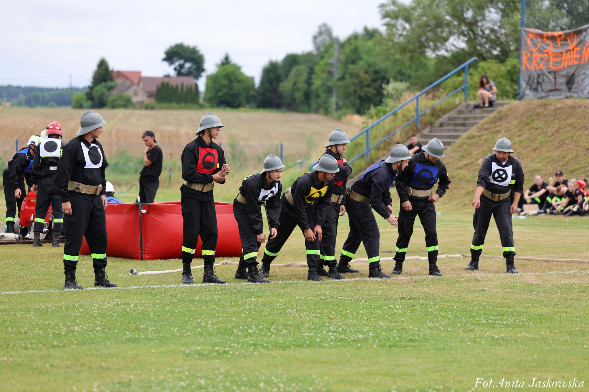 Mężczyźni w mundurach strażackich z hełmami i kolorowymi oznaczeniami na piersiach ustawieni na zielonym boisku obok czerwonego basenu i białego węża strażackiego.