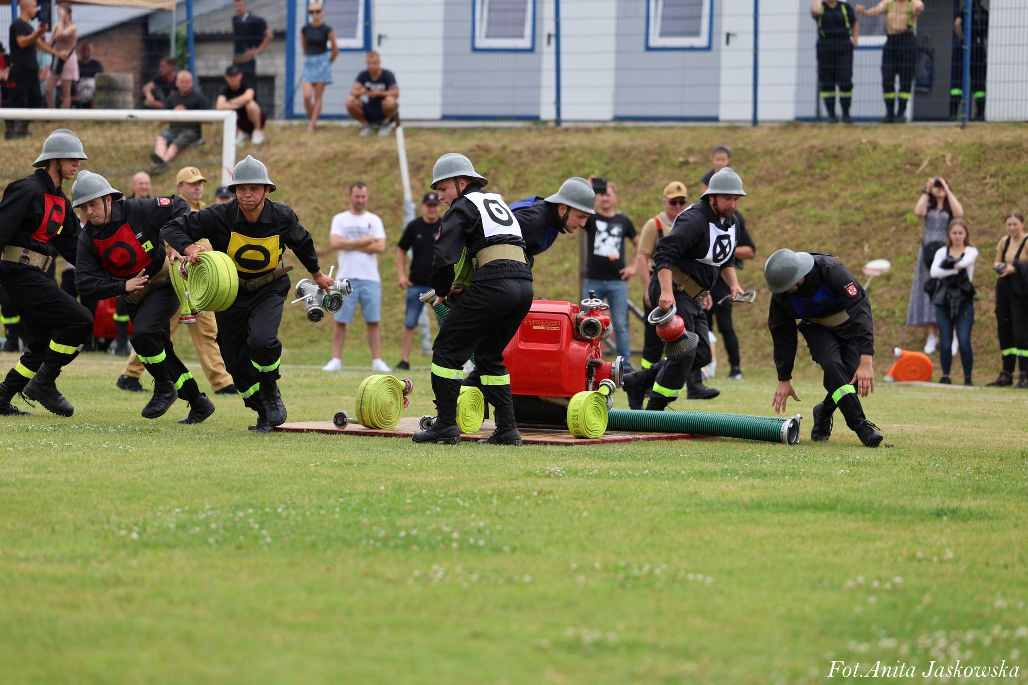 Grupa mężczyzn w hełmach i czarnych ubraniach z oznaczeniami, rozkładających zielone zwinięte węże i czerwony sprzęt na zielonym boisku, w tle widzowie.
