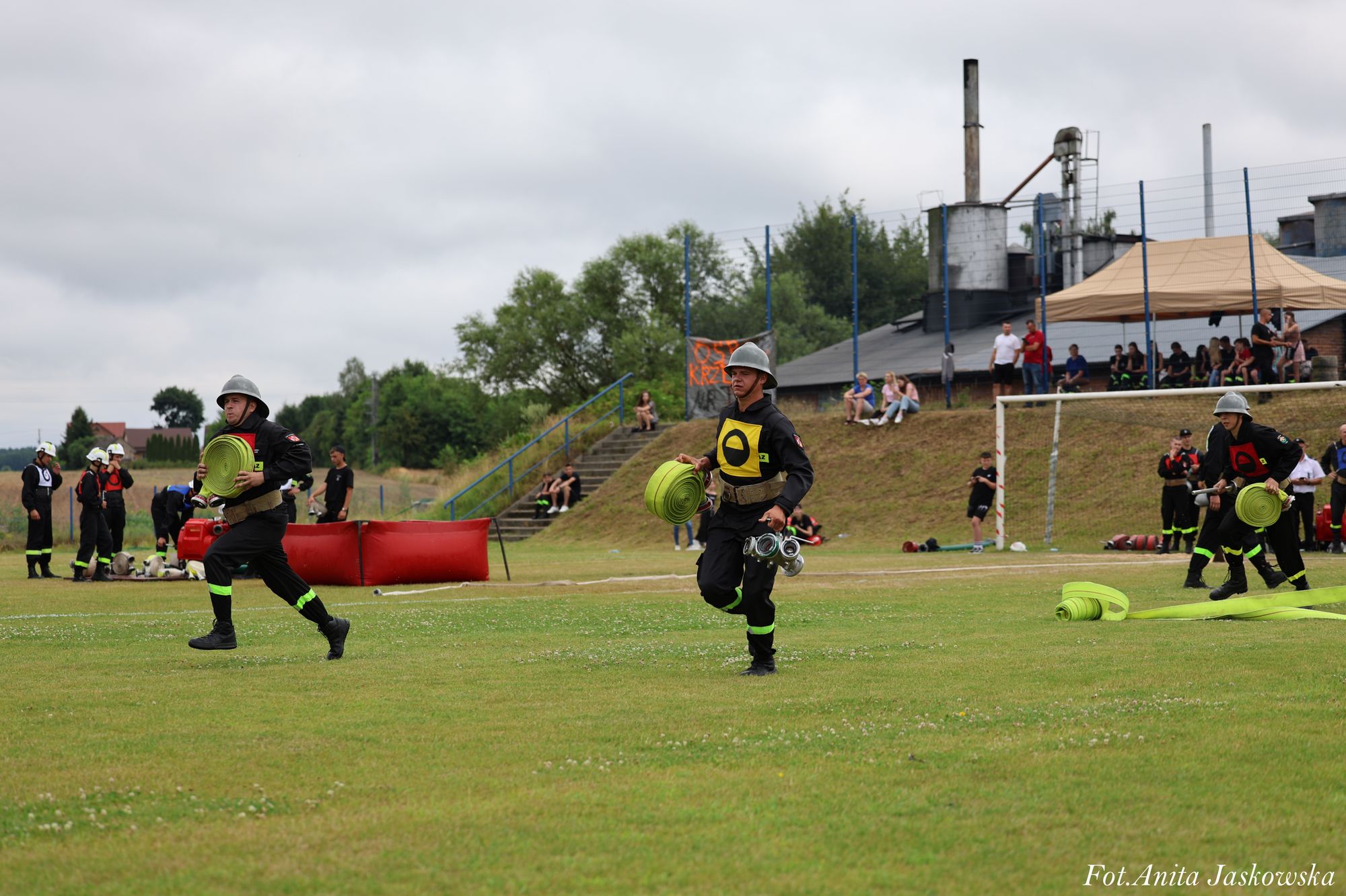 Strażacy w czarnych mundurach z odblaskowymi paskami i hełmach biegną po zielonym polu, niosąc zwinięte zielone węże strażackie, w tle ludzie na trybunach i budynki przemysłowe.