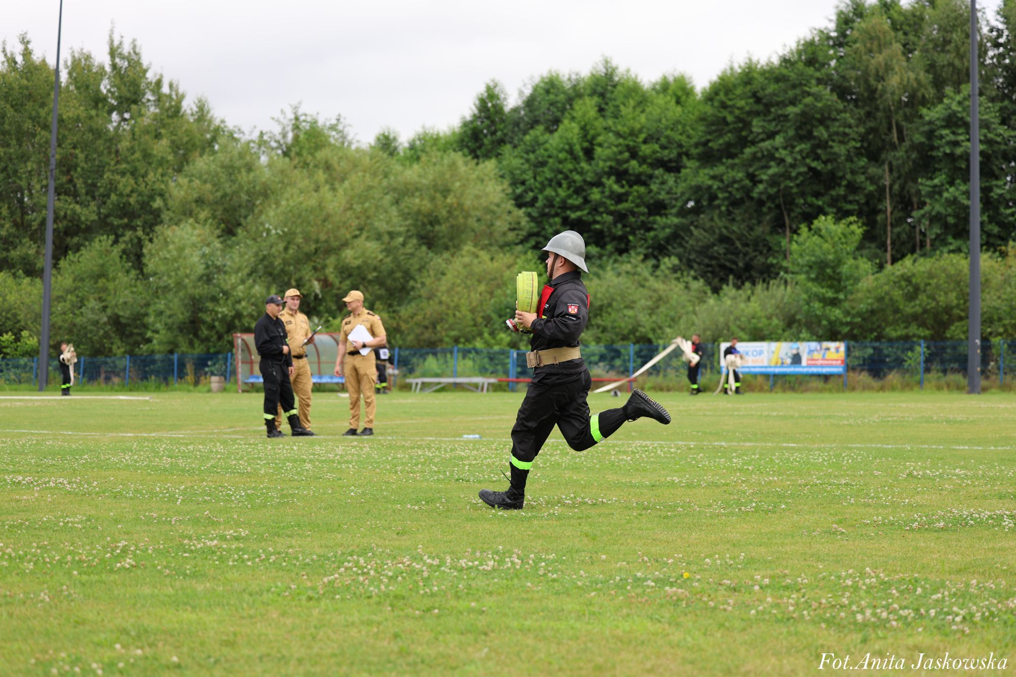 Mężczyzna w czarnym uniformie z neonowymi paskami biegnie po zielonym trawiastym boisku, trzymając zrolowany żółty wąż strażacki, w tle grupa osób w mundurach beżowych i czarnych.